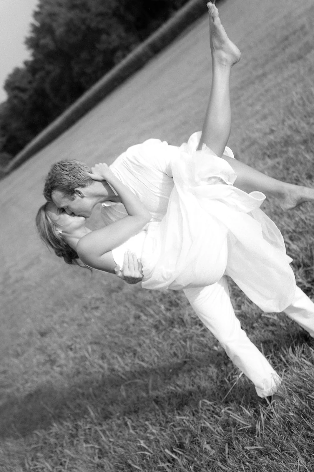 A black and white photo of a couple dressed in formal wedding attire, sharing a kiss outdoors on a grassy field with trees in the background. The man is holding the woman as they embrace.