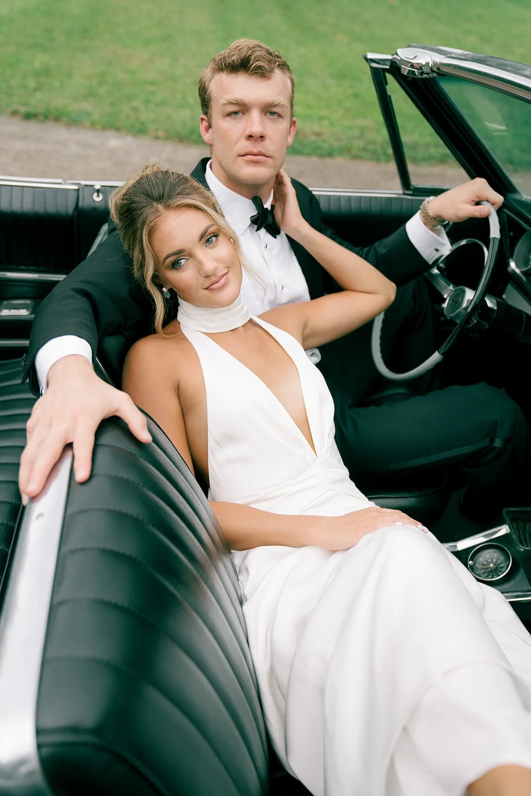 A couple dressed in formal wedding attire sitting in a vintage convertible car, with the woman resting her head on the man's shoulder.