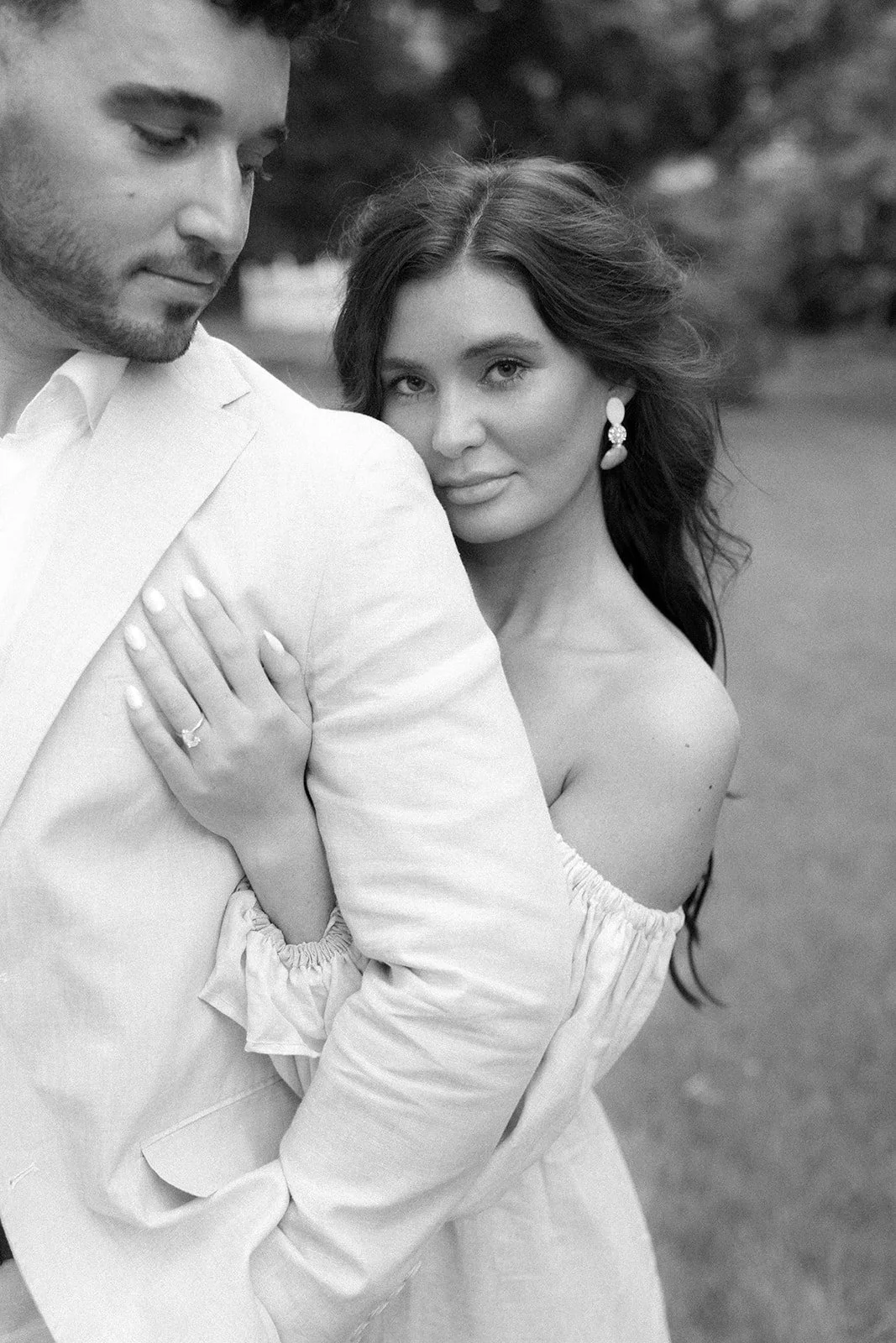 Black and white photo of a man and woman in an intimate pose outdoors, with the woman leaning against the man's shoulder, looking at the camera, and the man facing downward. The woman has long hair, earrings, and an off-the-shoulder top, while the ma