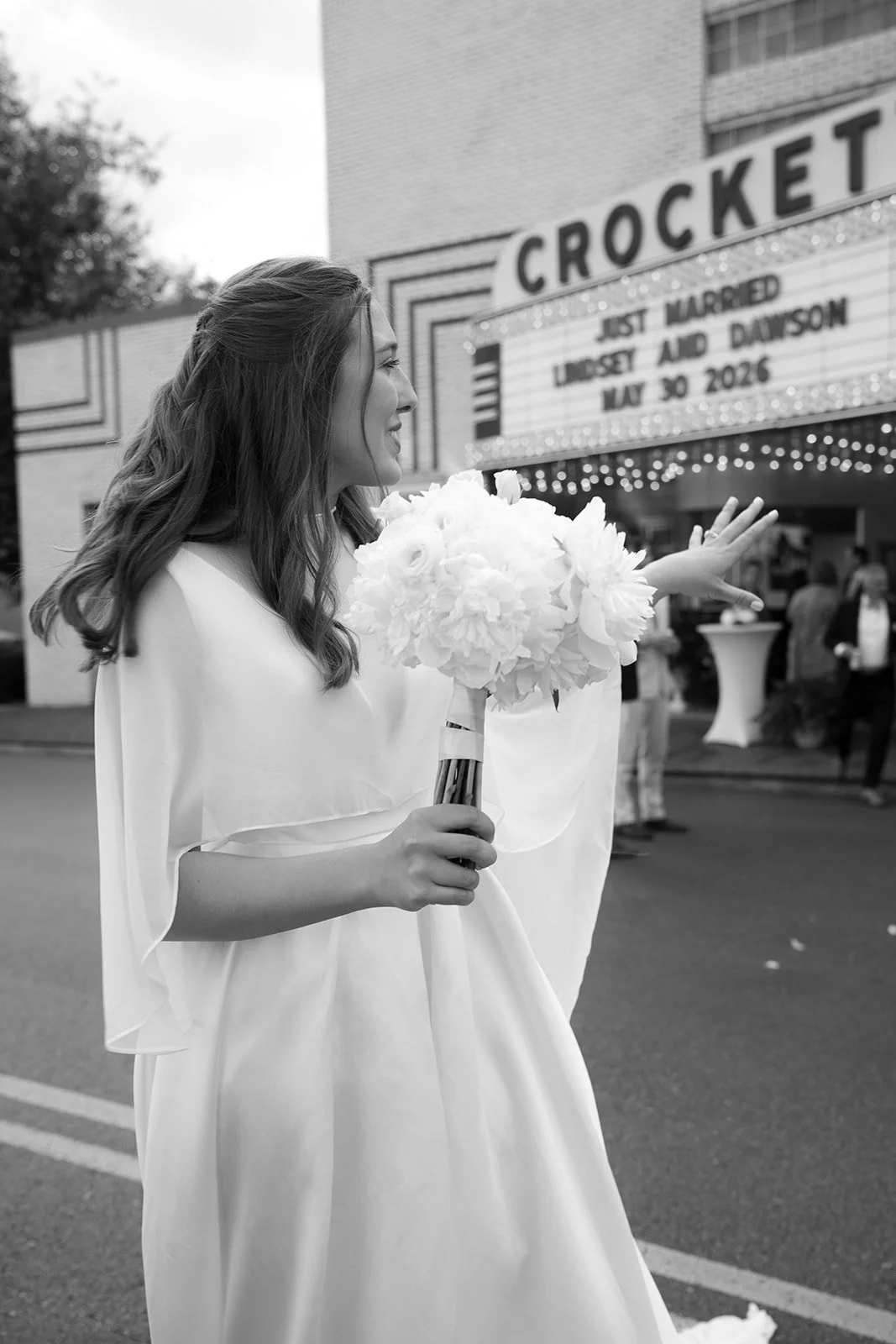 A woman in a wedding dress holding a bouquet of flowers, smiling, standing outside a theater with a marquee sign that reads 'Just Married Lindsay and Dawson May 30 2026'.