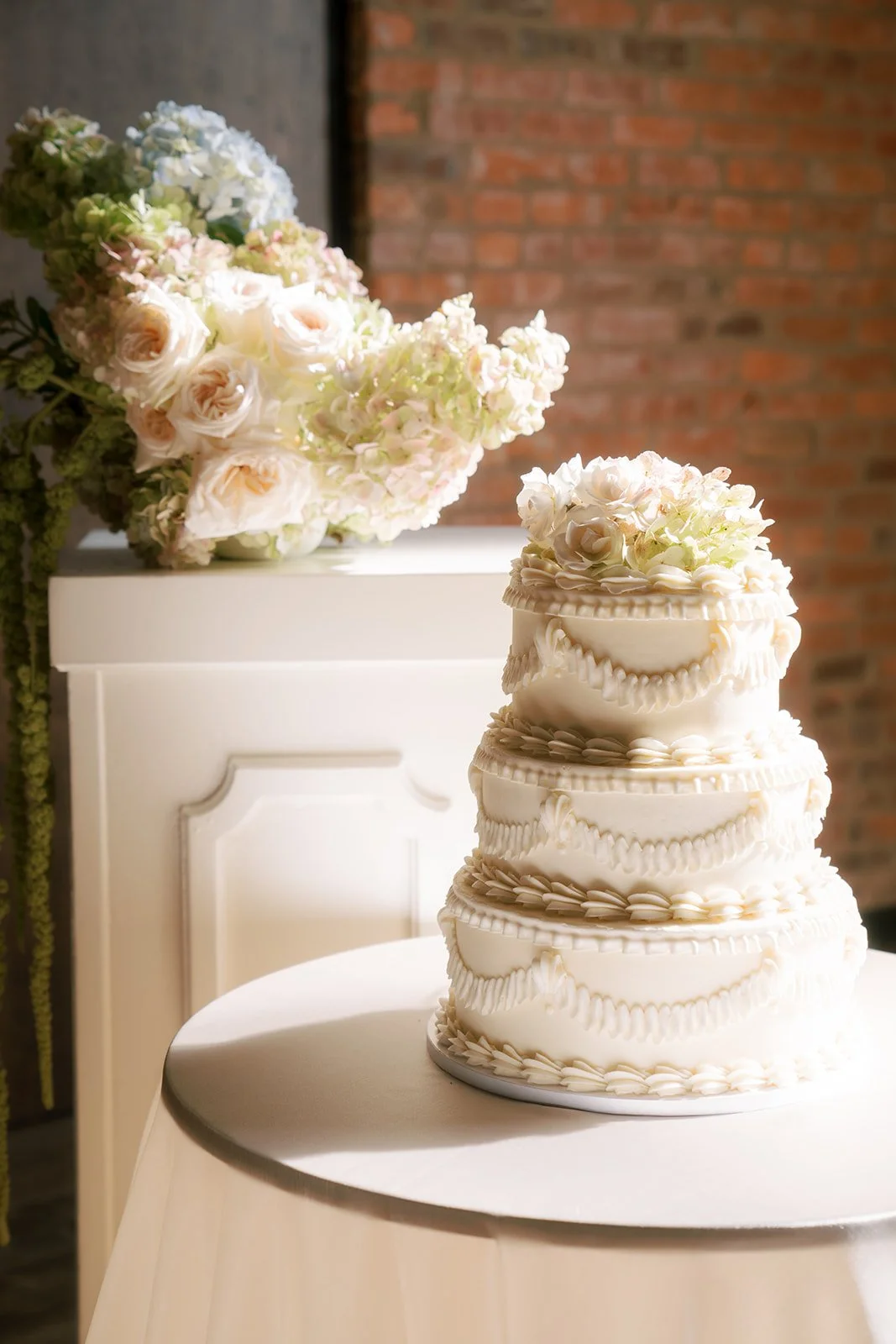 A multi-tier white wedding cake decorated with white flowers and cream piping, placed on a round table with a floral arrangement of white, pink, and blue flowers beside it. Background features a brick wall and a white furniture piece.