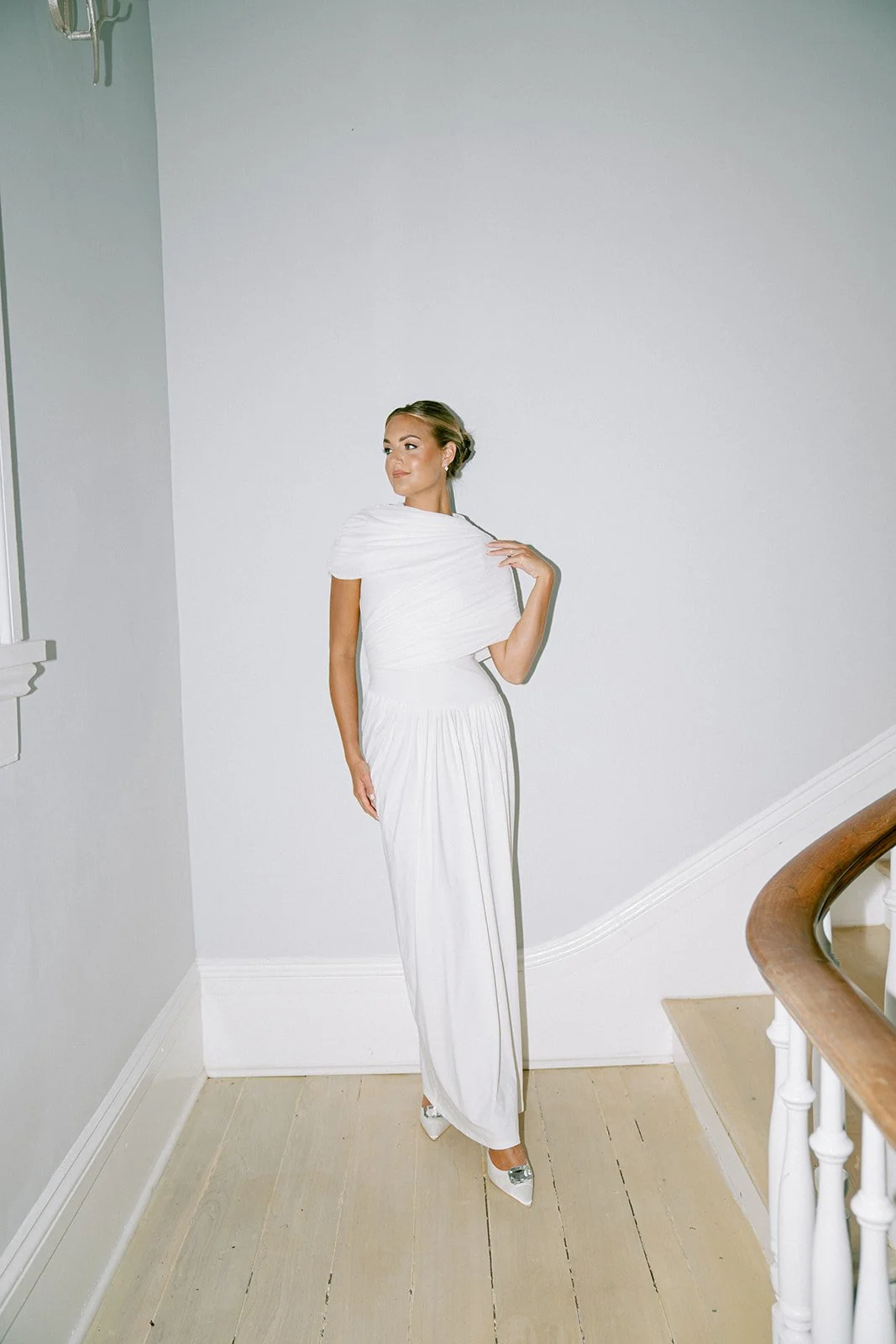 A woman in a white gown standing on a wooden floor next to a staircase, posing against a plain white wall, with her hair styled in an elegant updo.
