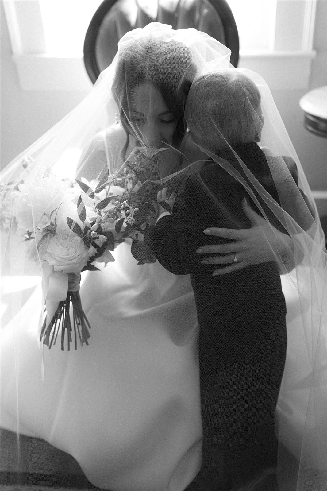 A bride wearing a veil and holding a bouquet of flowers hugs a young girl.