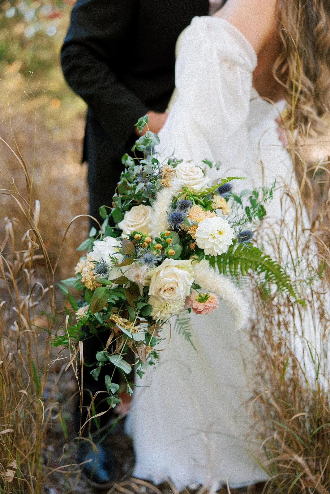 Bride holding a cascading bouquet of white roses, cream-colored flowers, ferns, and greenery in a natural outdoor setting with tall dried grass around.