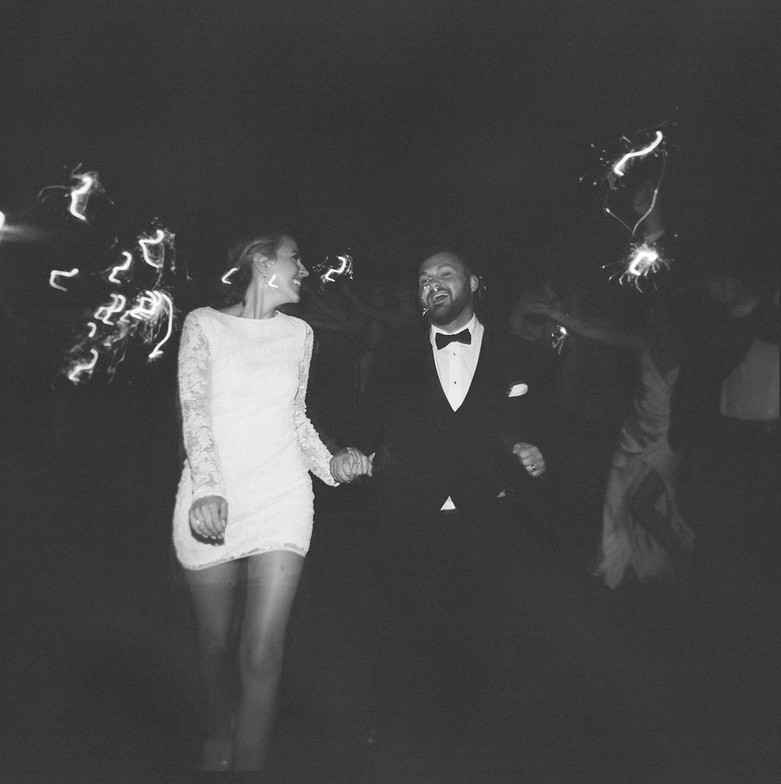 A black and white photo of a bride and groom holding hands and smiling, standing on a dance floor during their wedding reception. The bride wears a long white lace wedding dress and the groom is dressed in a tuxedo with a bowtie. There are sparklers burning behind them.