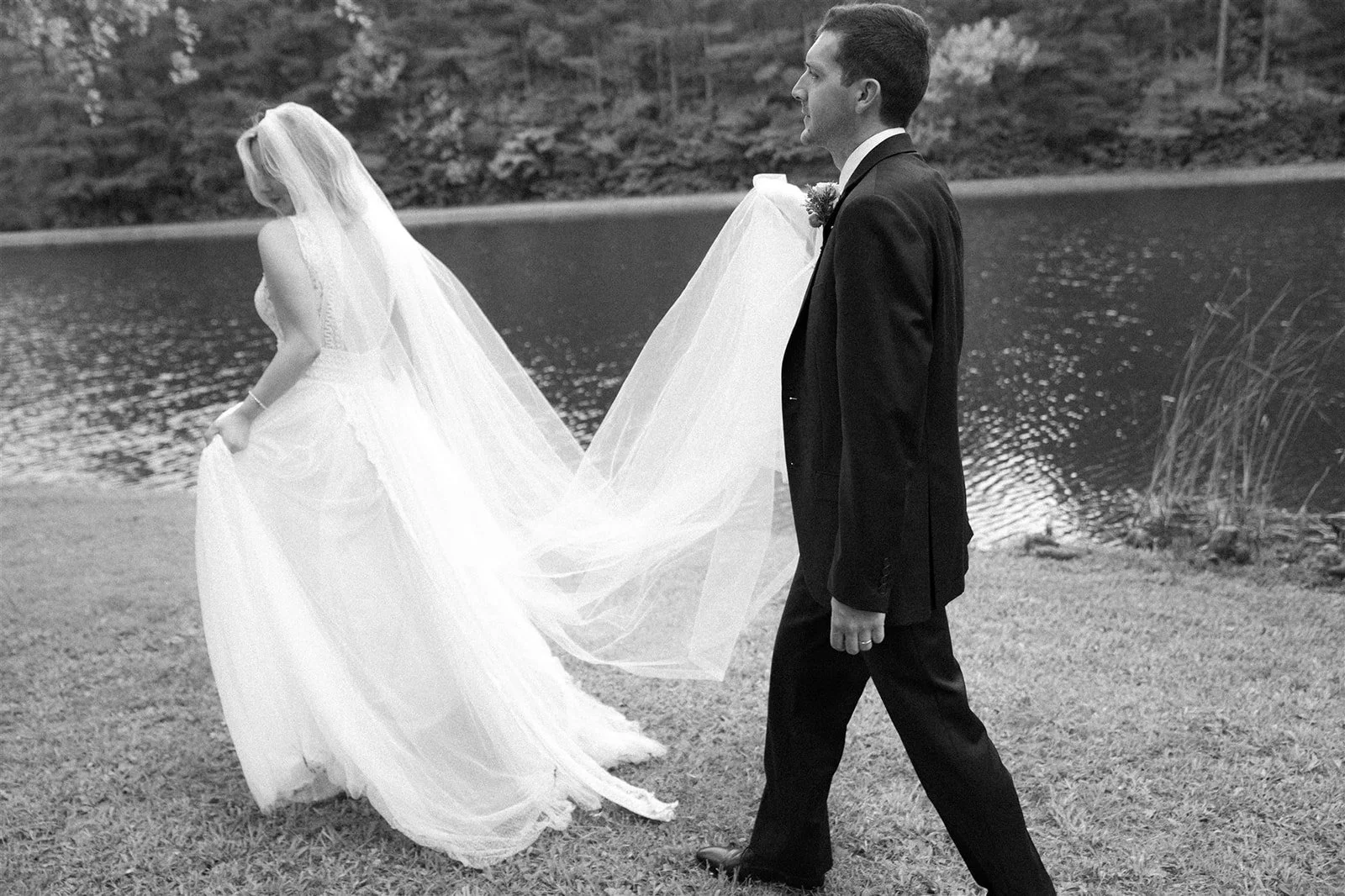 A black-and-white photo of a bride and groom walking outdoors by a lake. The bride is holding her wedding dress and the groom is smiling, holding a bridal veil that connects to the bride's dress. There are trees in the background.