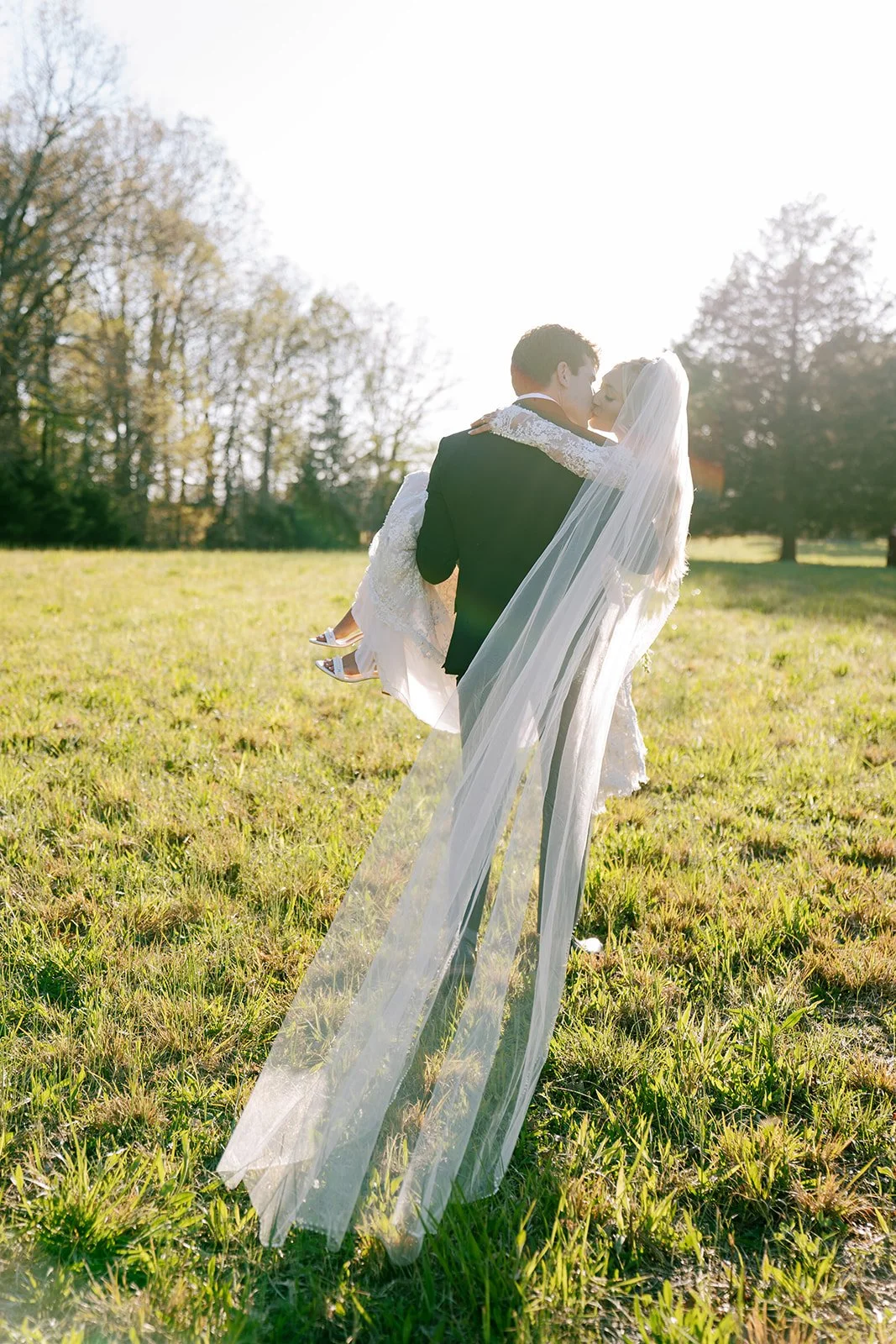 A bride and groom in wedding attire outdoors during daytime. The groom is carrying the bride, who is wearing a veil and holding her dress, in a grassy field with trees in the background.