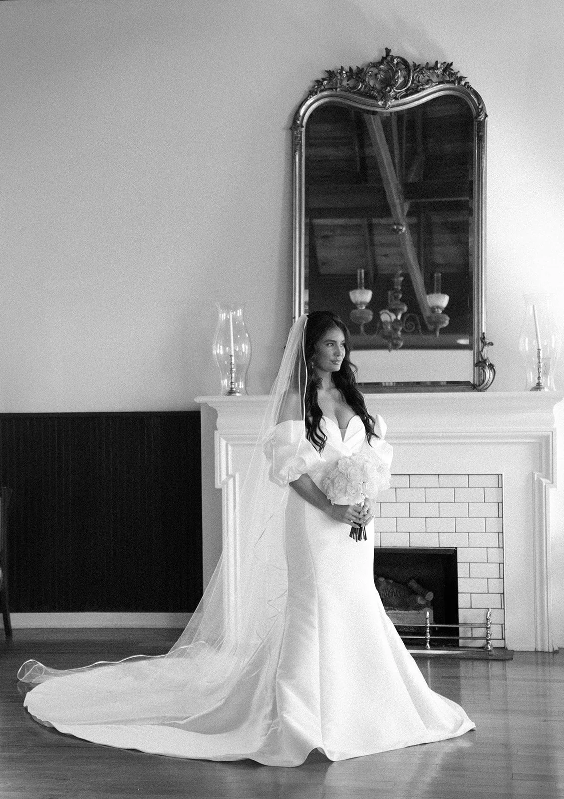 A bride in a wedding gown holding a bouquet standing in front of a fireplace with a large ornate mirror above.