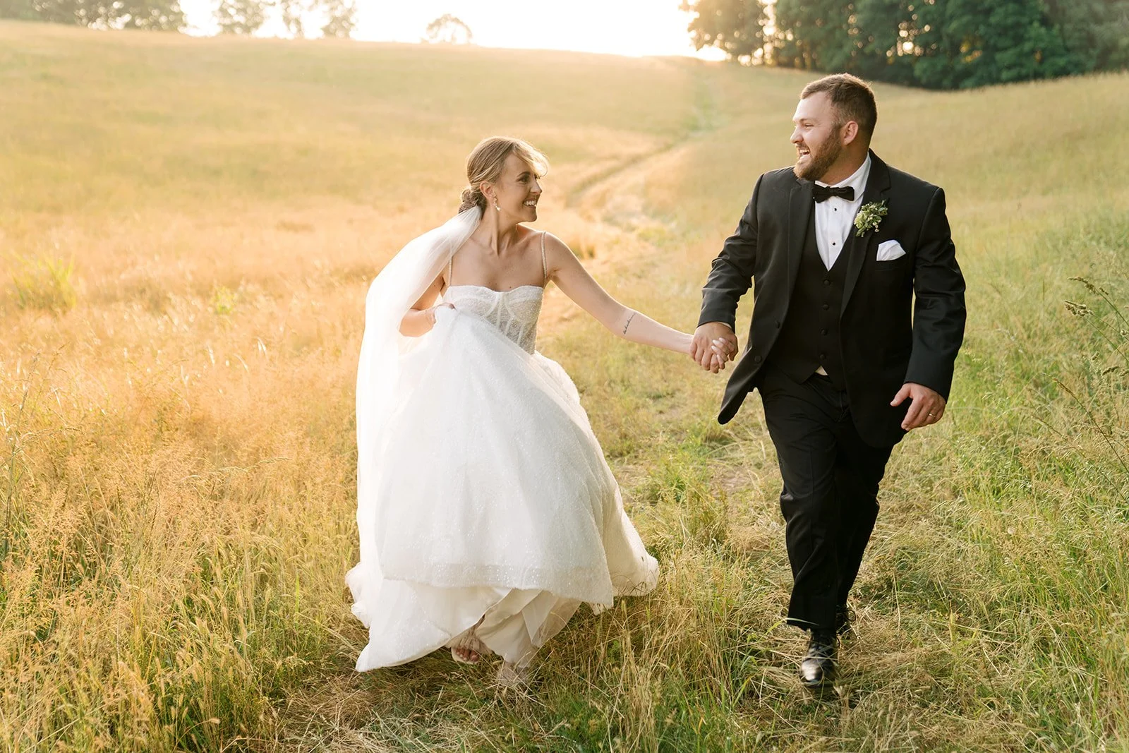 A newlywed couple walking hand in hand through a grassy field at sunset, smiling and looking at each other, with trees in the background.