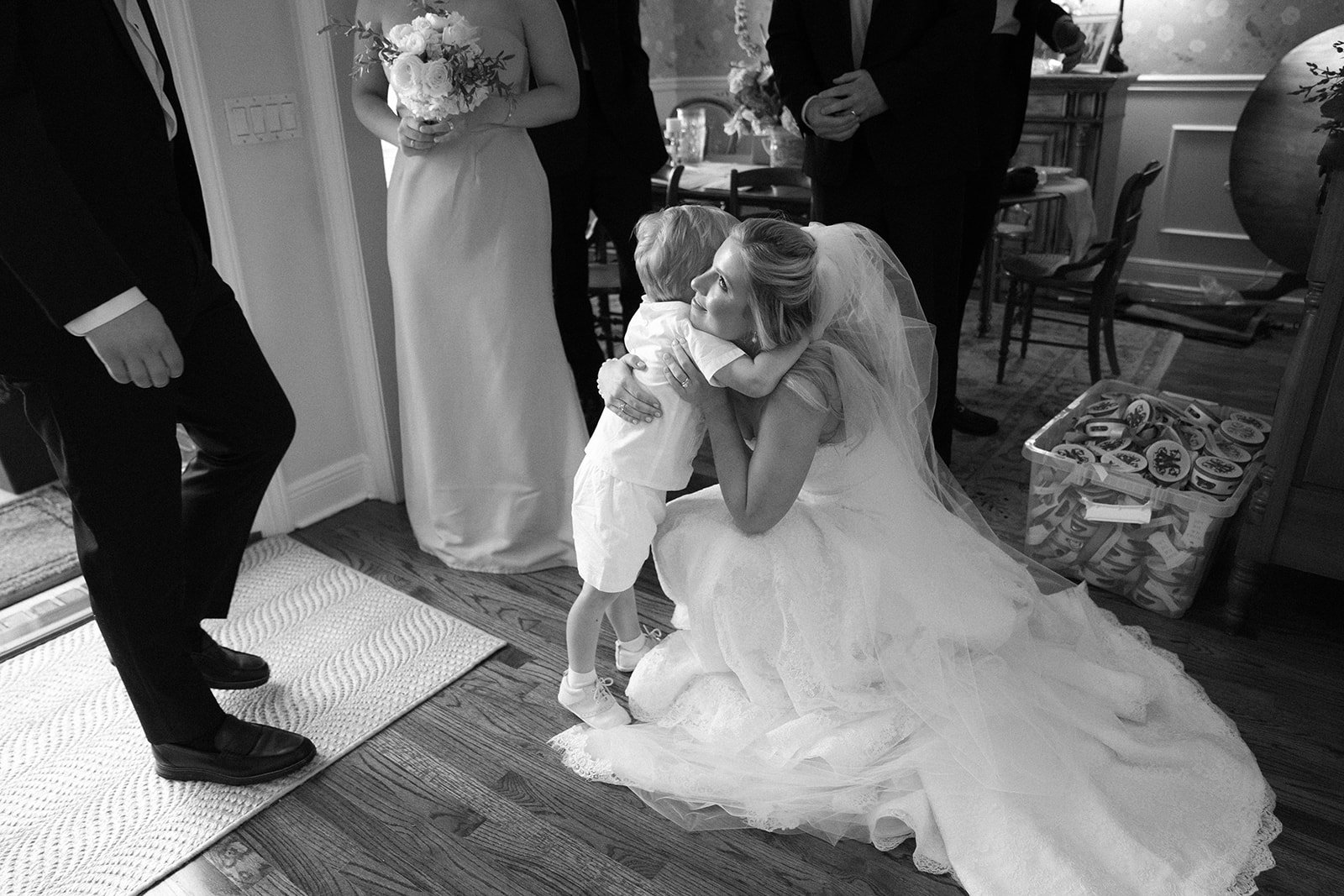 A bride kneeling and hugging a small child at a wedding reception, with wedding guests standing nearby, in a decorated room.