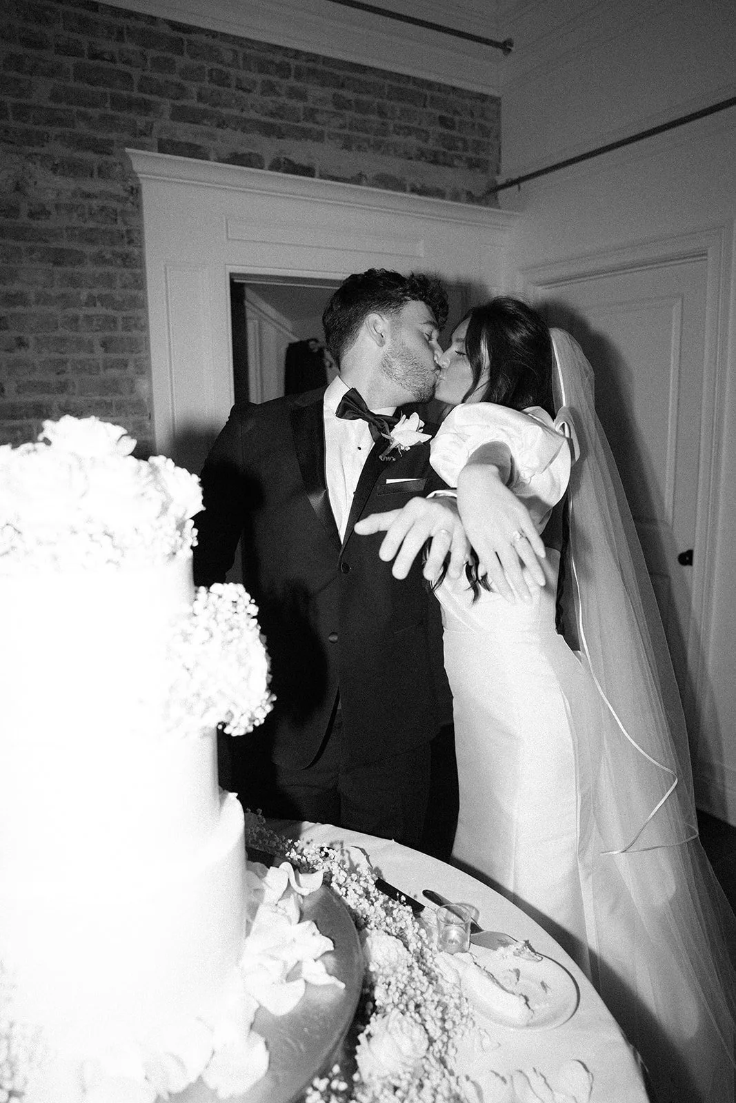A bride and groom share a kiss at their wedding, dressed in formal attire, with wedding cake and desserts on a table in the foreground.