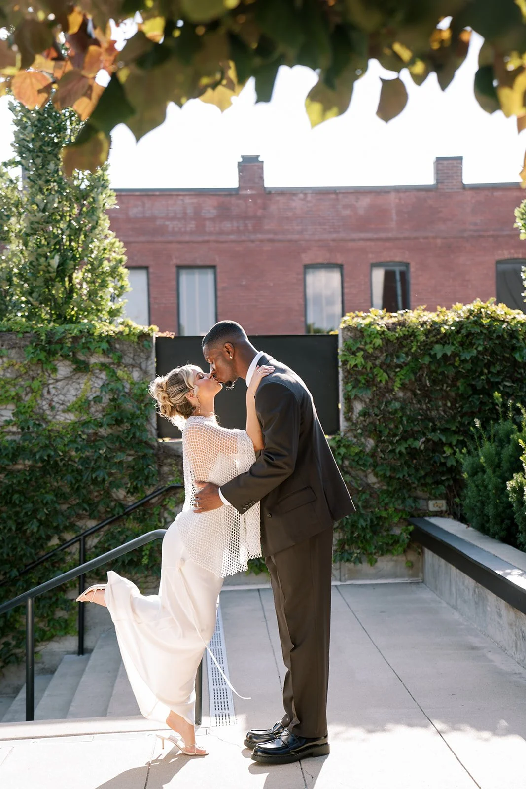 A couple sharing a kiss outdoors, with the woman in a white dress and the man in a black suit, surrounded by greenery and brick buildings in the background.