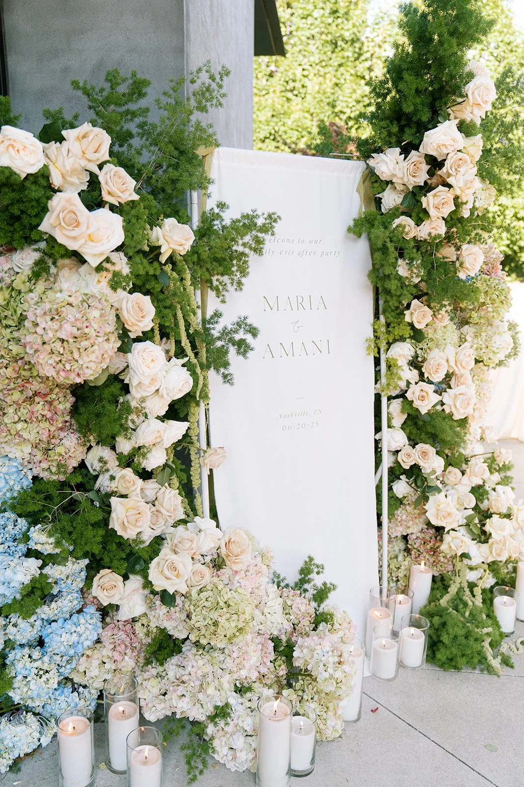 Wedding sign with white backdrop surrounded by pastel-colored hydrangeas, roses, and greenery, with candles at the base, sign reads 'Welcome to our happily ever after party' with names Maria and Amani and date June 20, 2023.