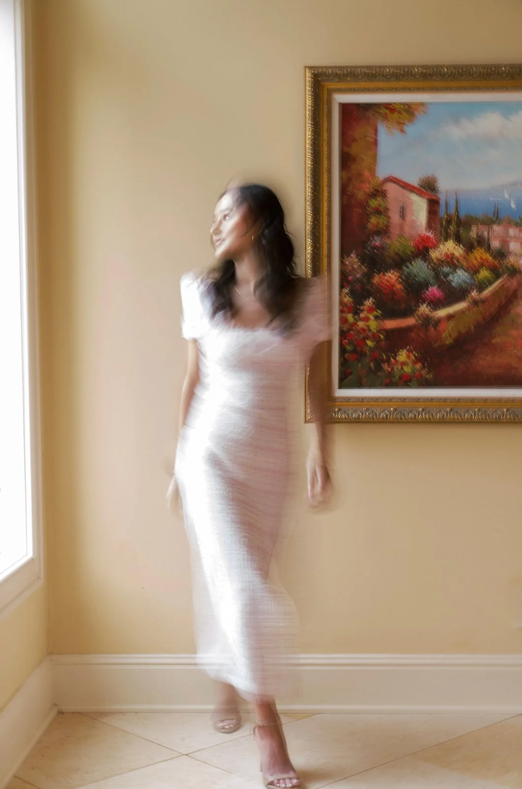 Blurry image of a woman in a white dress standing next to a framed painting of a colorful landscape on a yellow wall, near a window.