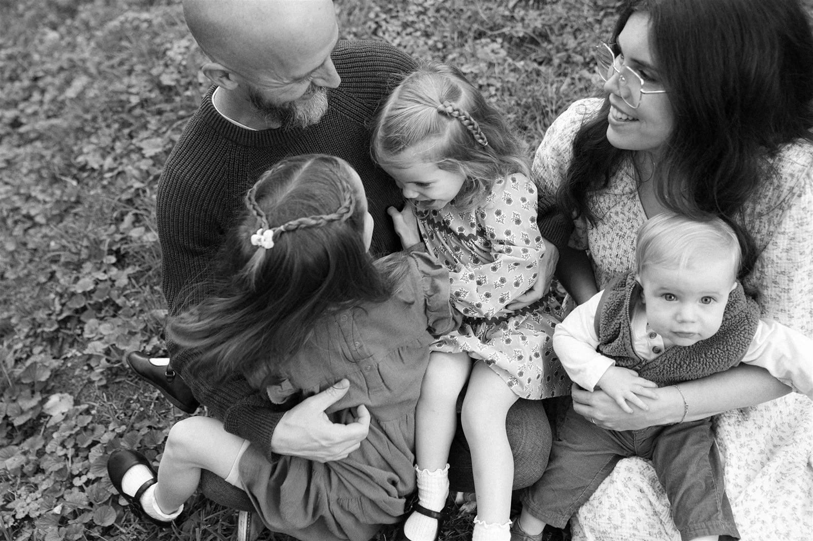 A black-and-white photo of a happy family sitting outdoors on grass, with two adults and three children, engaged in an affectionate moment.