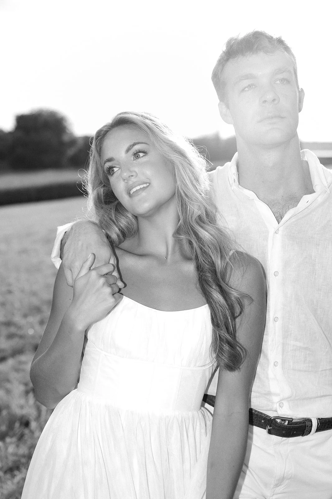 A black-and-white photo of a young woman and a young man outdoors. The woman has long wavy hair and is wearing a sleeveless white dress, looking slightly to her left with a gentle smile. The man, standing behind her with his arm around her shoulder, 