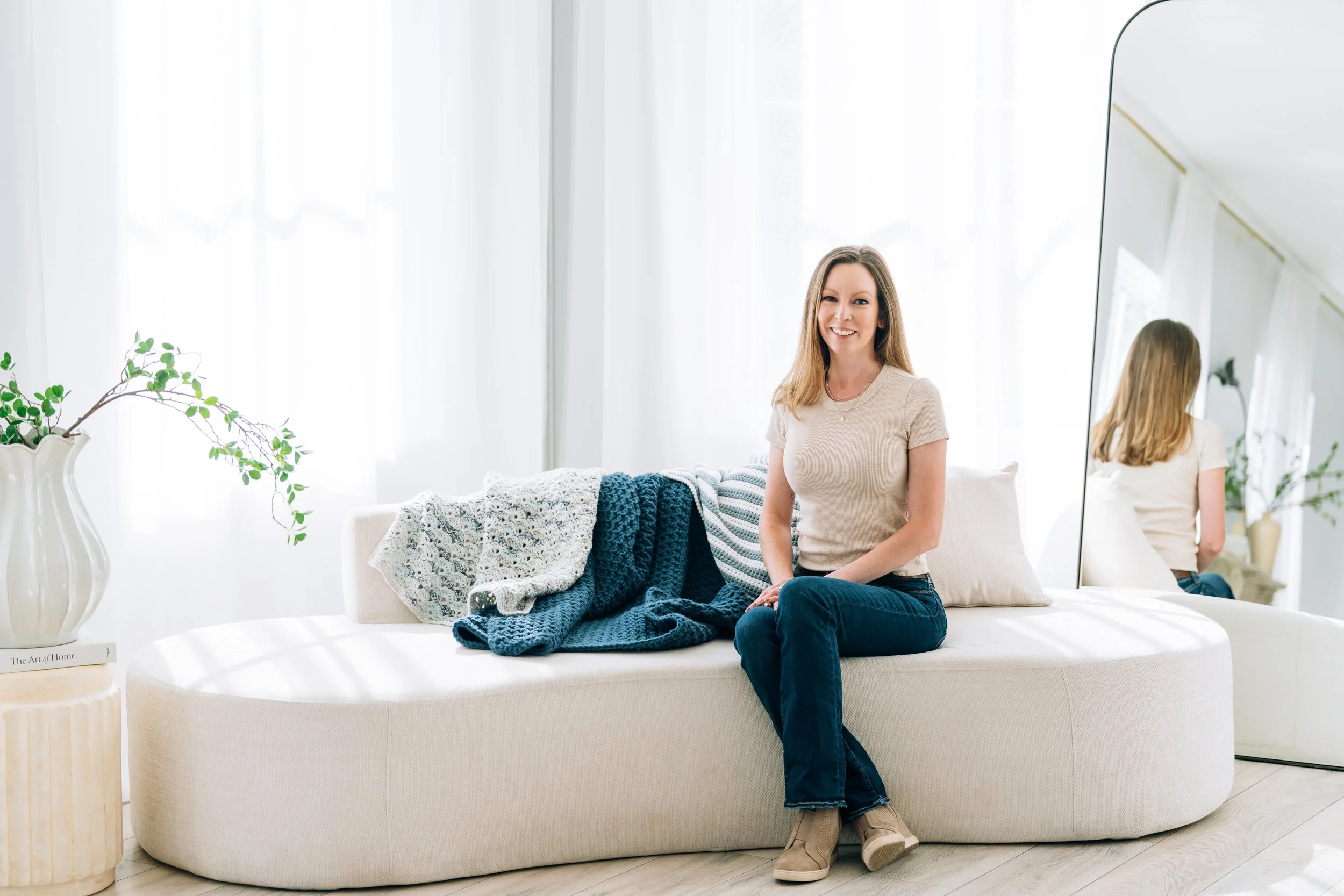 A woman sitting on a white sofa in a bright, airy room with sheer white curtains, a large mirror, and a decorative plant in a vase on a small table.