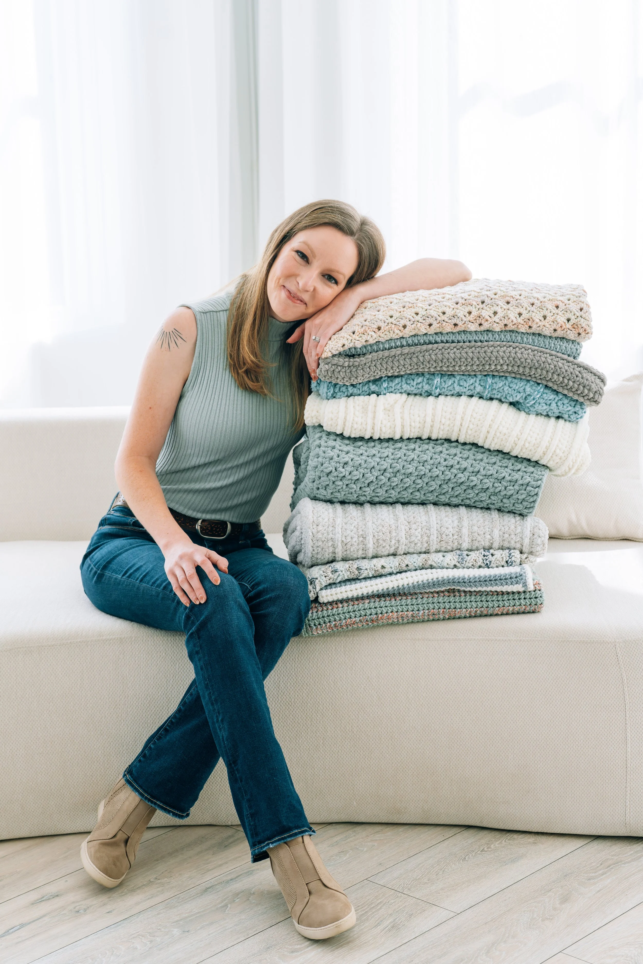 A woman sitting on a beige sofa, leaning on a tall stack of folded knitted blankets in neutral and pastel colors, in a bright room with white curtains.