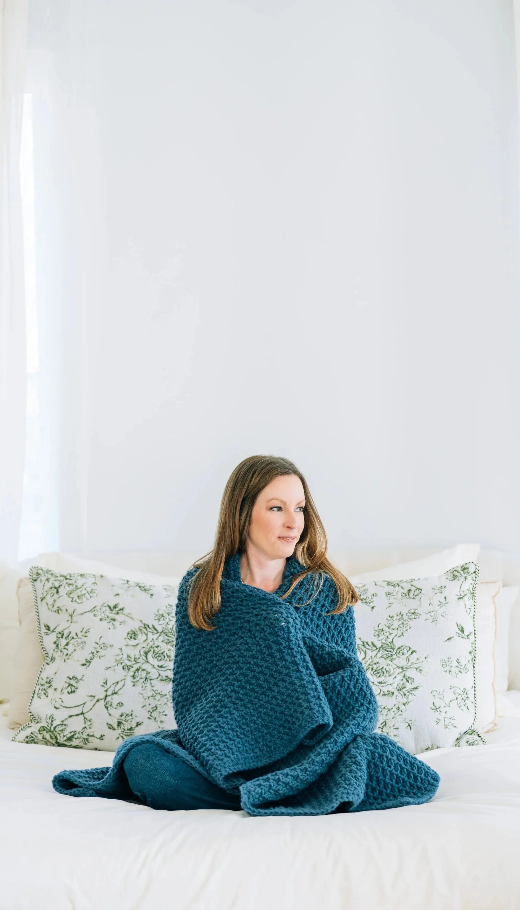 A woman with long brown hair, wearing a blue knitted sweater, sits on a bed with white sheets and pillows that have green floral patterns, in a room with white walls and a window.