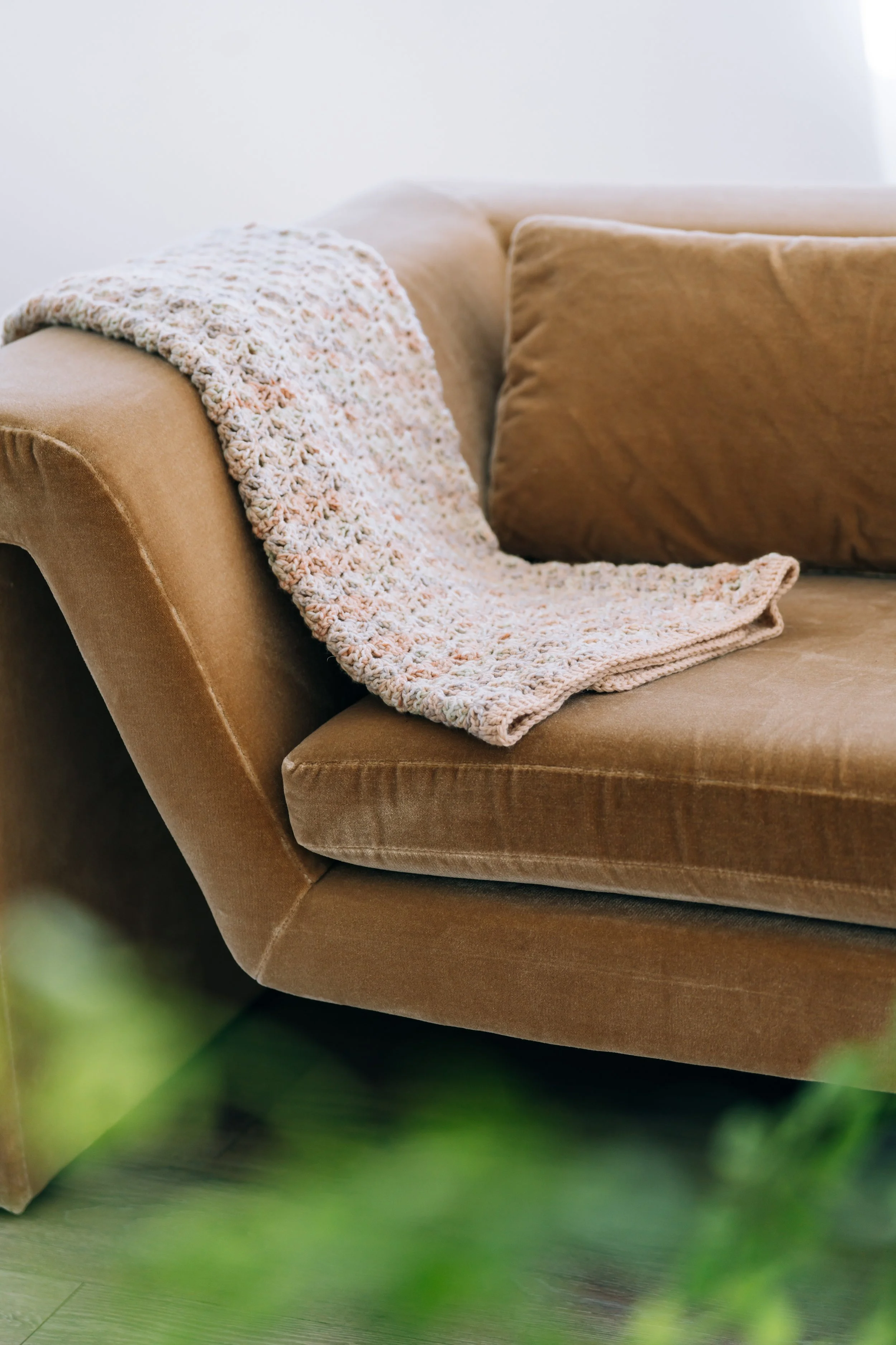 Close-up of a beige sofa with a crocheted throw blanket draped over the armrest and a mustard-colored cushion on the backrest, with some greenery blurred in the foreground.