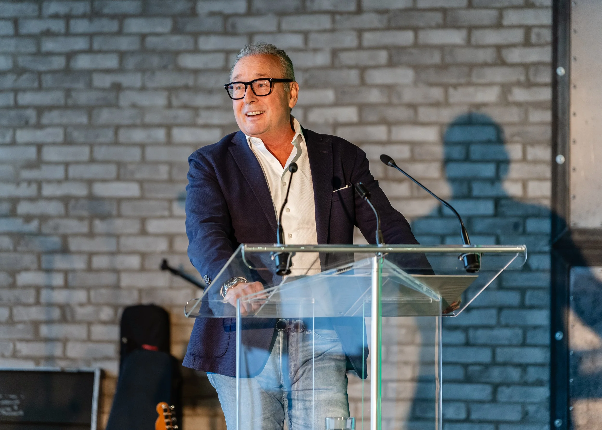 A man with gray hair, wearing glasses, a dark blazer, and a white shirt, is standing at a clear lectern with microphones, smiling while speaking. The background is a brick wall.