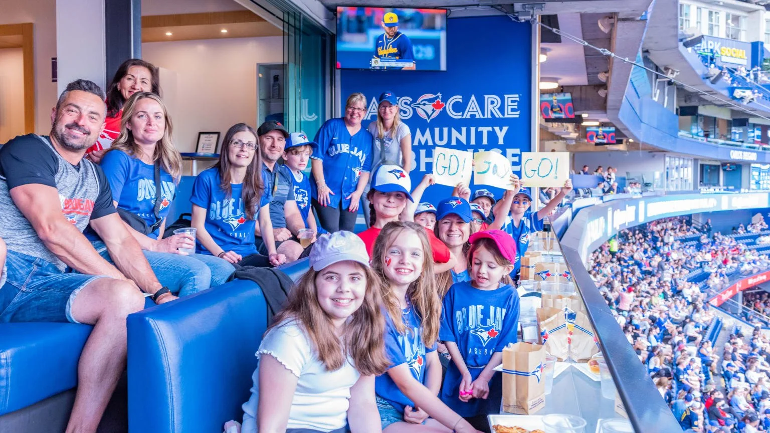 Family and friends cheering at a baseball game in the stadium, some holding signs saying 'G.O. G.O. G.O.' and wearing Toronto Blue Jays apparel.