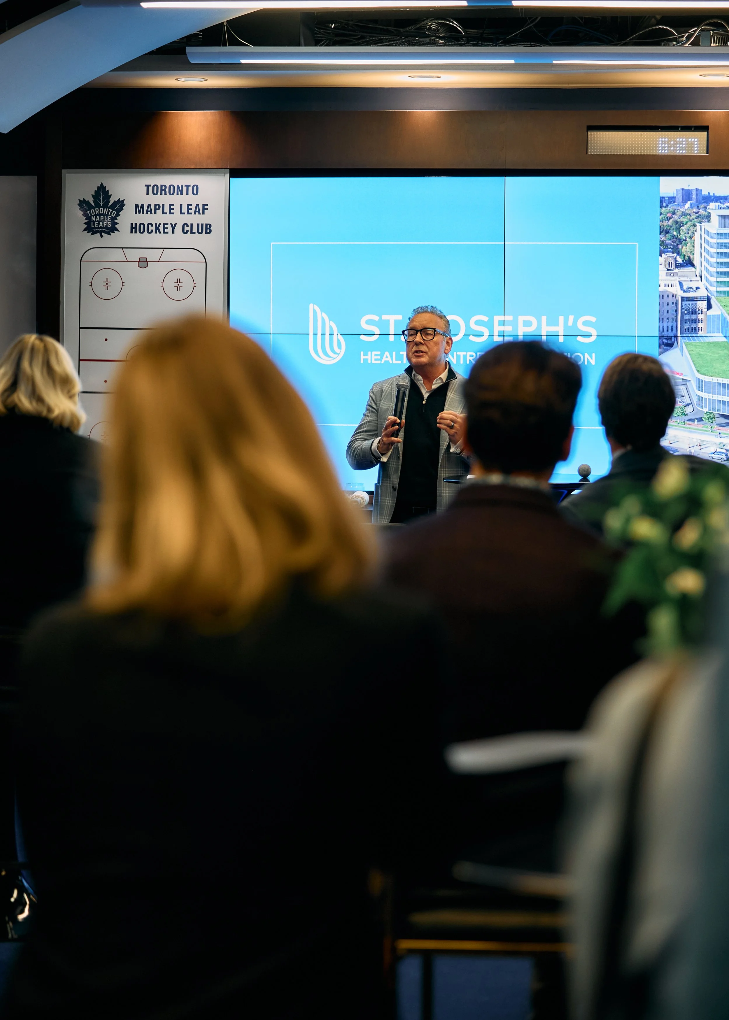 A man giving a presentation in a conference room with a large screen displaying the St. Joseph's Health Centre Foundation logo and a cityscape. The audience is seated, and a Toronto Maple Leaf logo with hockey schematic is visible on the wall.