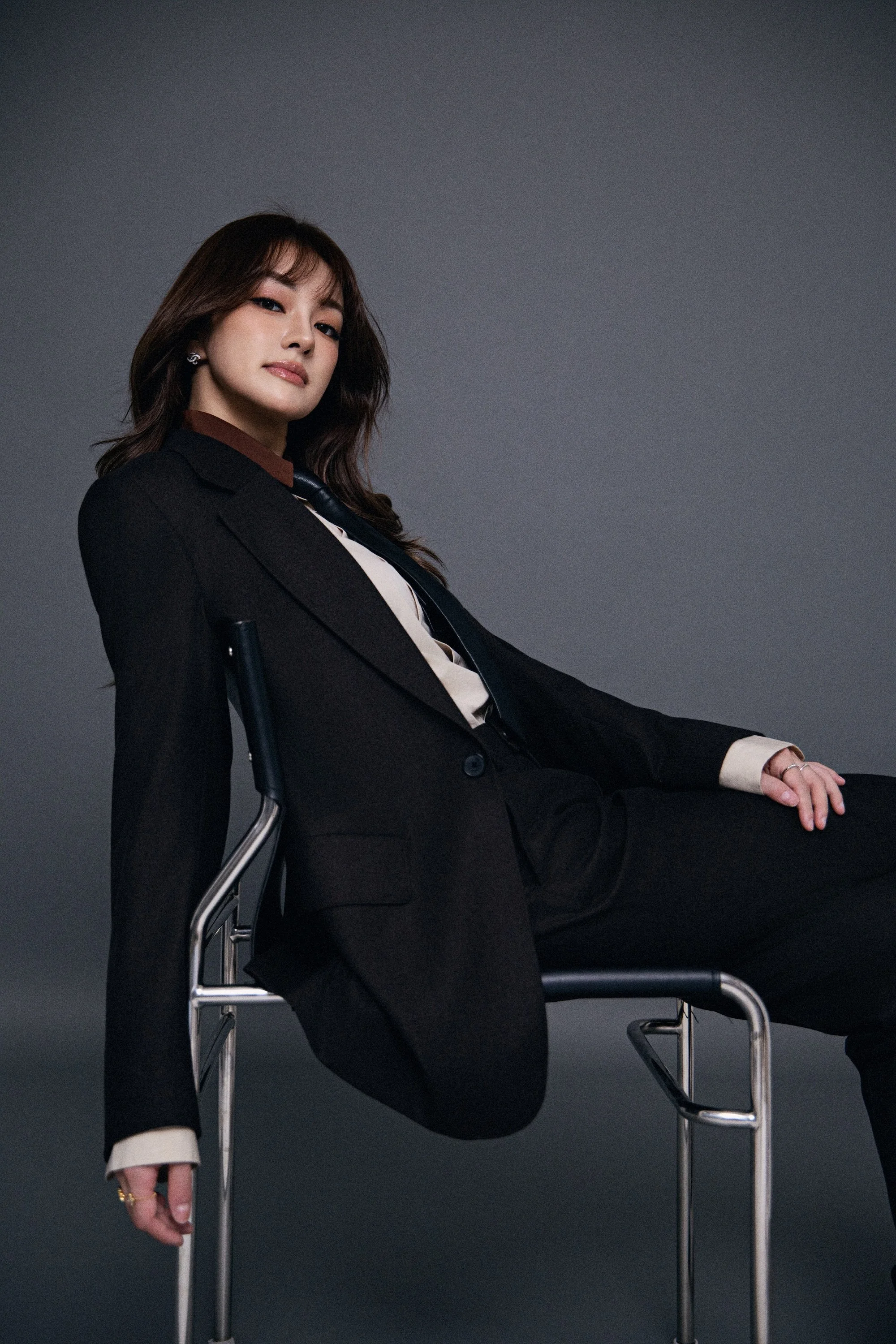 A woman dressed in a black blazer and pants, sitting on a metal chair with one leg crossed over the other, posing against a dark gray background.