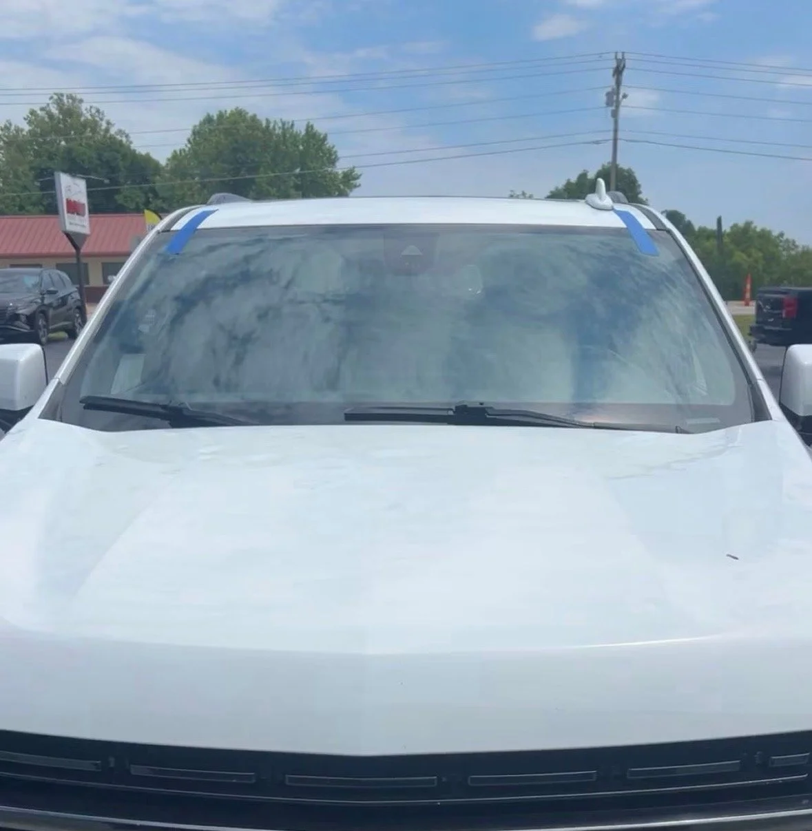 Front view of a white car with blue tape on the windshield and the top edges of the vehicle, parked outdoors with other vehicles, trees, and power lines in the background.
