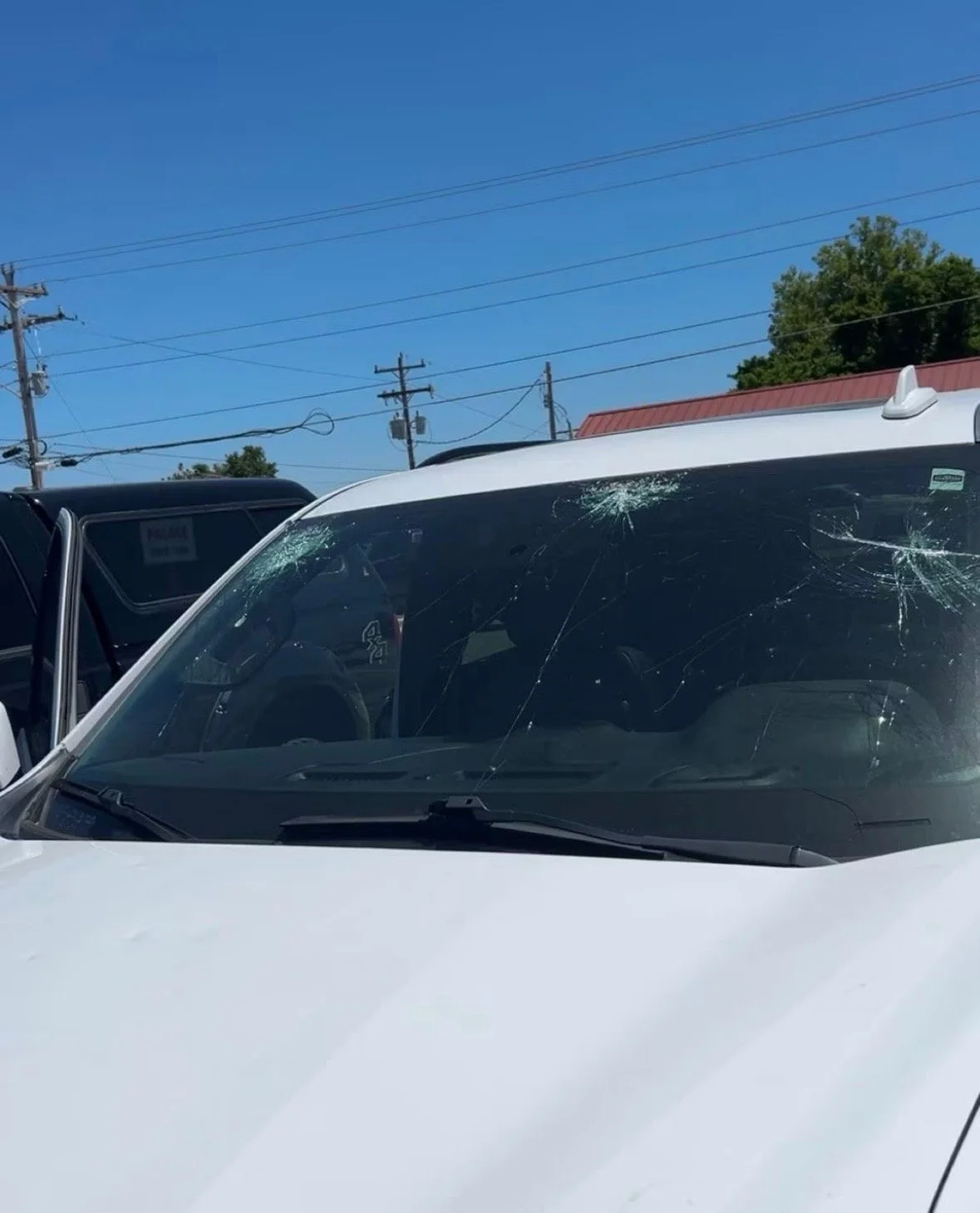 White vehicle with multiple cracks and shattered glass on the windshield, parked outdoors under a clear blue sky with utility poles in the background.