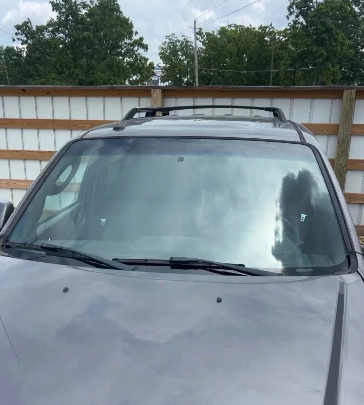 Front of a gray SUV with a roof rack parked in front of a white wooden fence and green trees, with cloudy sky reflected on the windshield.