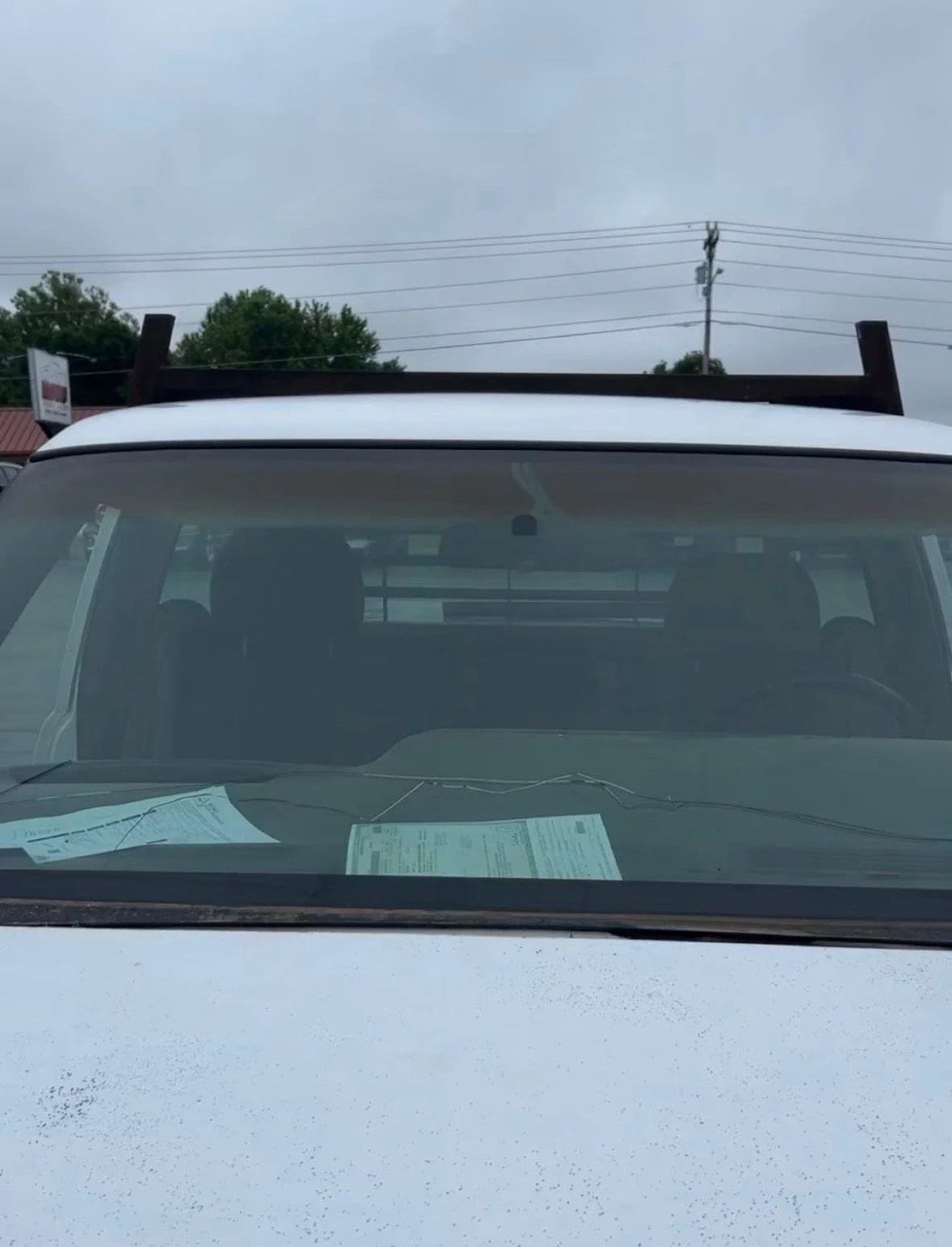 The front view of a white vehicle with a roof rack, seen through the windshield with paperwork on the dashboard. Overhead, power lines and trees are visible under a cloudy sky.
