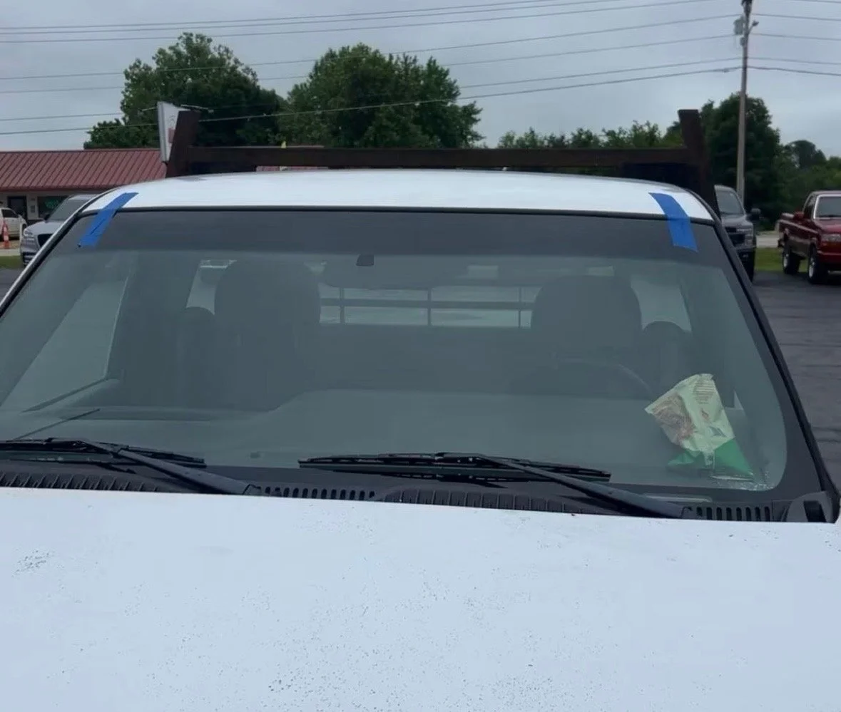 Front view of a white pickup truck with a brown ladder rack mounted on the bed, parked in a lot with other vehicles and trees in the background.