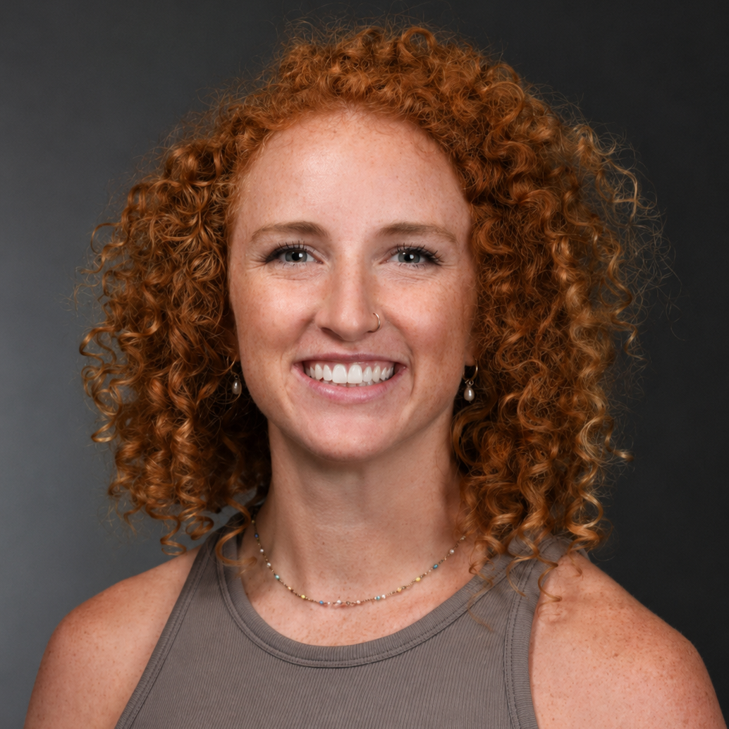 Portrait of a smiling woman with curly red hair, freckles, wearing a sleeveless gray top, earrings, and a necklace, against a dark background.