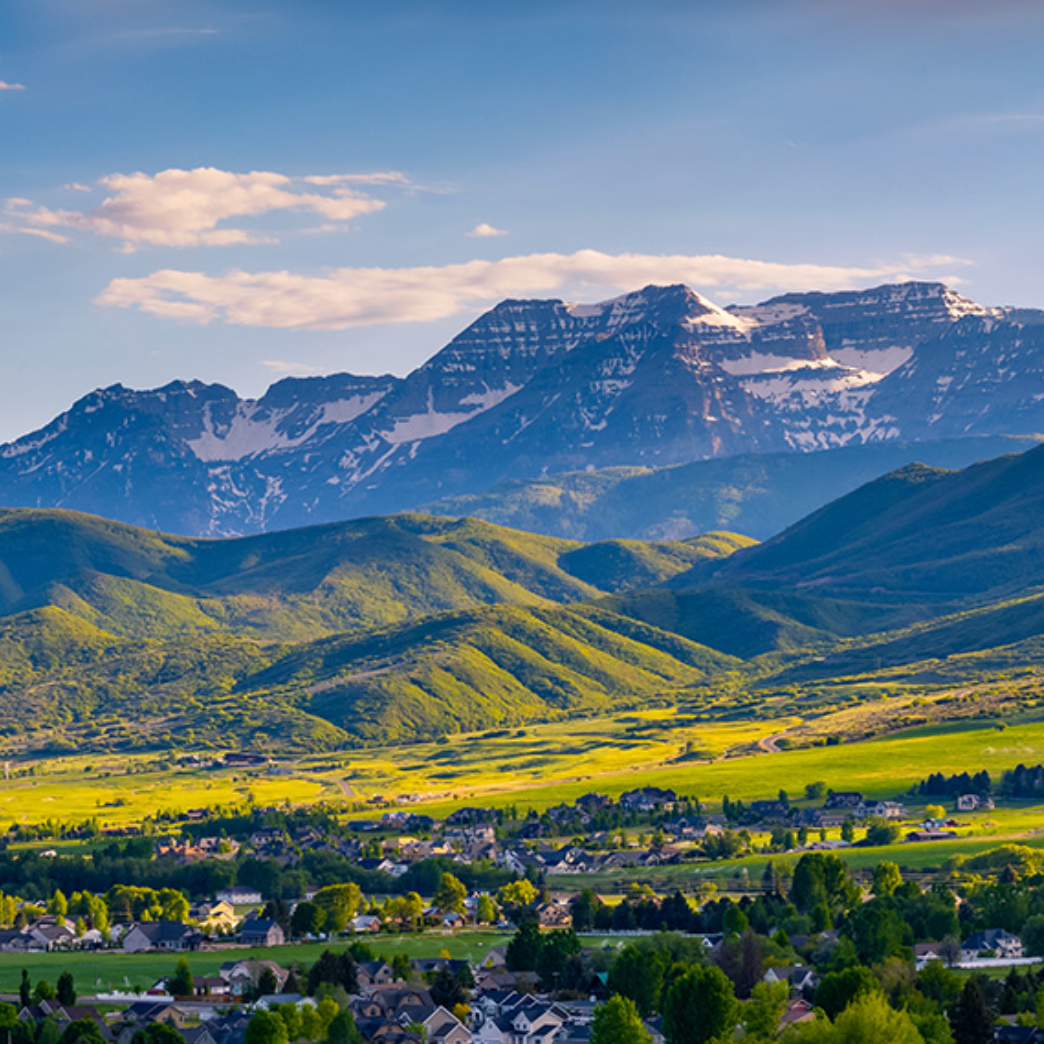 Scenic view of snow-capped mountains in the background, rolling green hills in the middle ground, and a small town with houses and trees in the foreground.