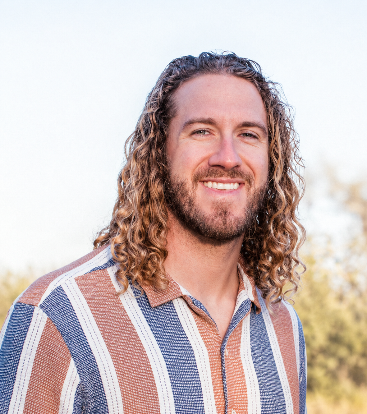 A man with long, curly hair and a beard smiling outdoors.