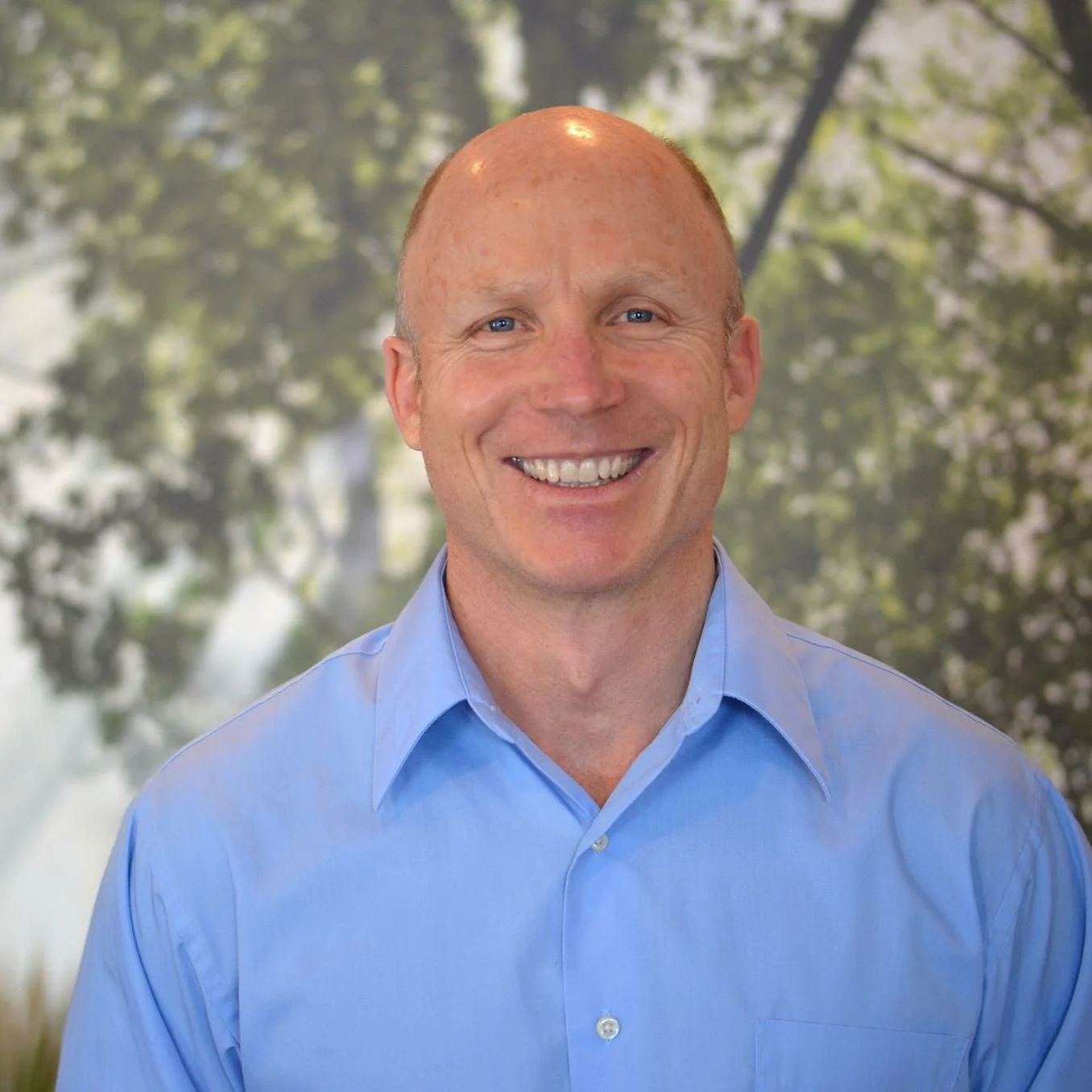 A smiling man with blue eyes and a bald head wearing a light blue shirt, standing outdoors with blurred trees in the background.