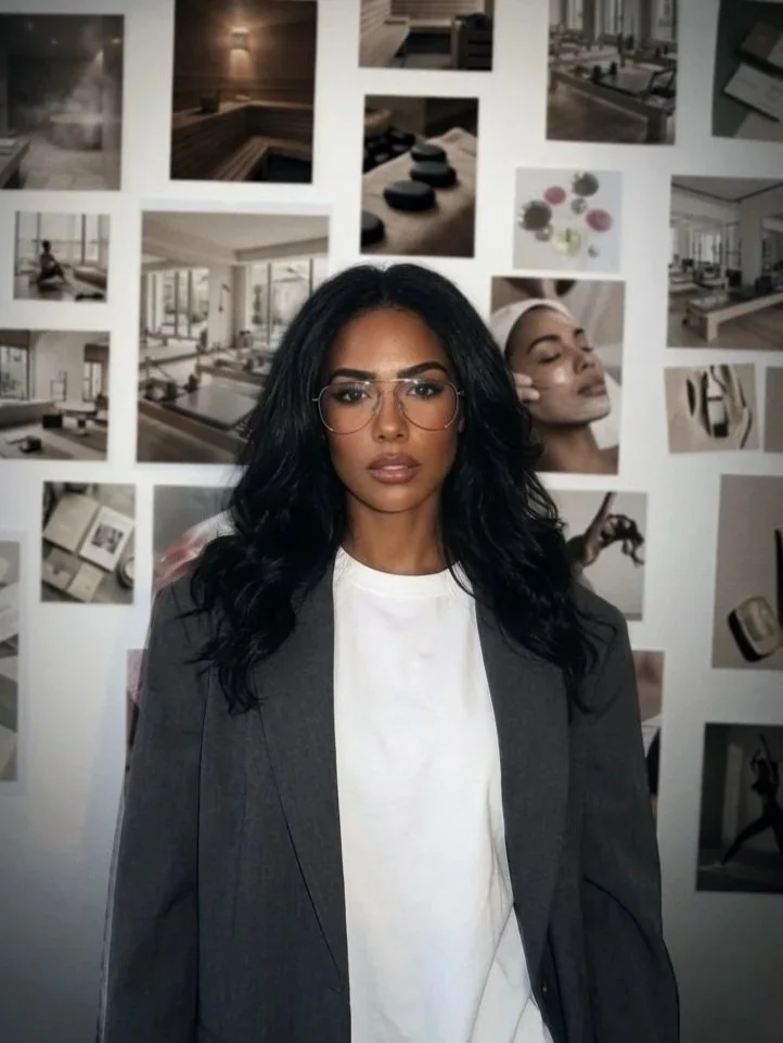 A woman with long dark hair, wearing glasses, a white t-shirt, and a dark blazer, standing in front of a wall of photographs, some of which feature interior design and objects.