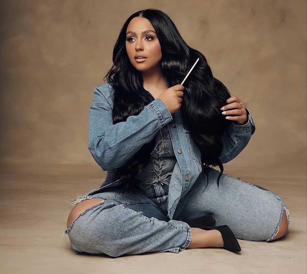 A woman sitting on the floor with long black hair, wearing a denim jacket and ripped jeans, holding a grooming brush.