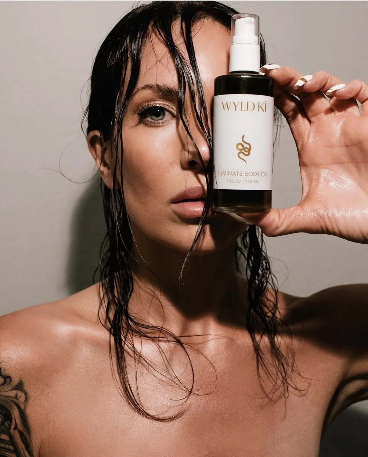 A woman with wet hair holding a bottle of Wyldki Illuminate Body Oil in front of her face, revealing her eye and lips, against a plain background.