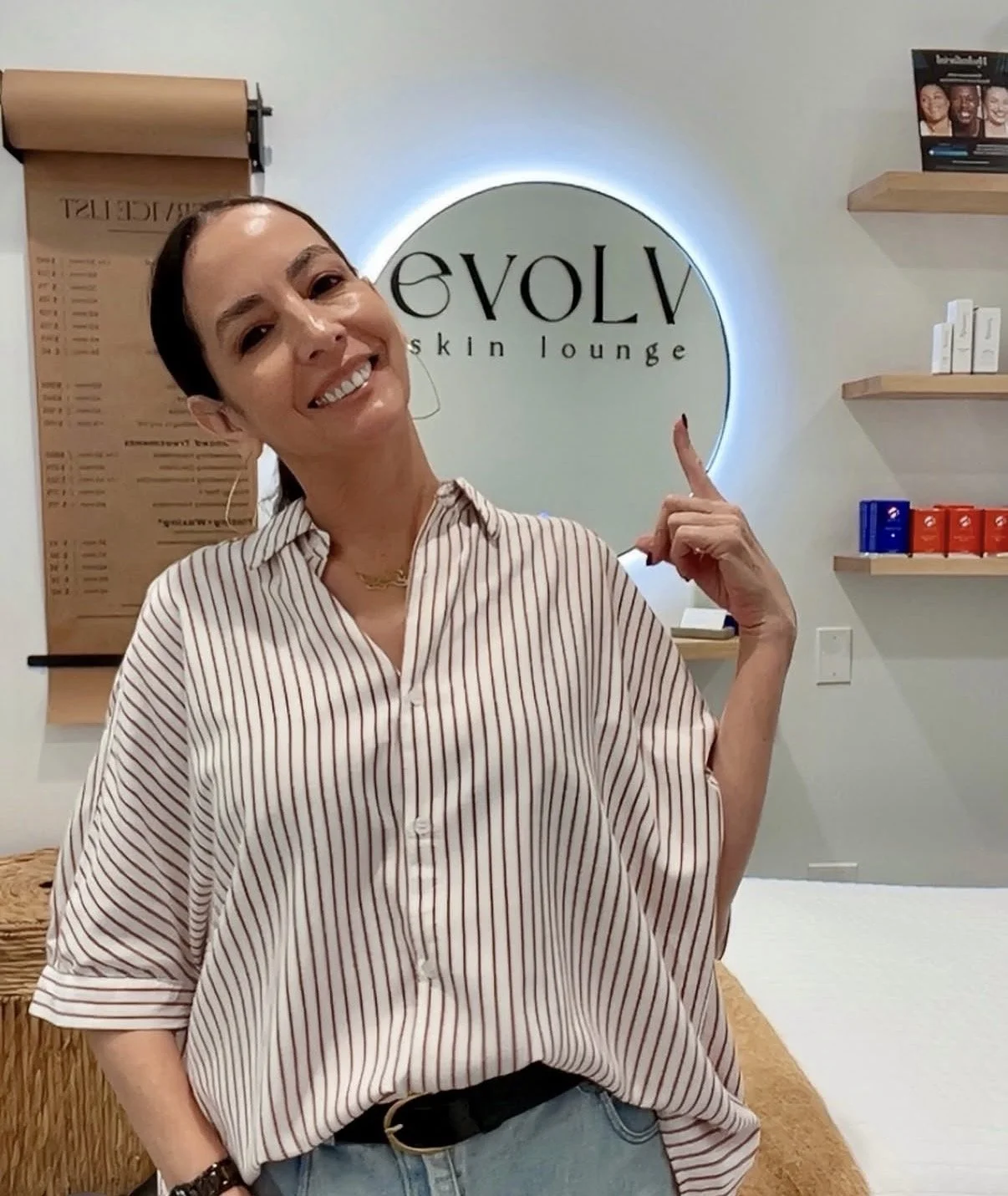 A woman smiling and pointing at a sign that says "EVOLVE skin lounge" in a wellness spa or skincare lounge interior.