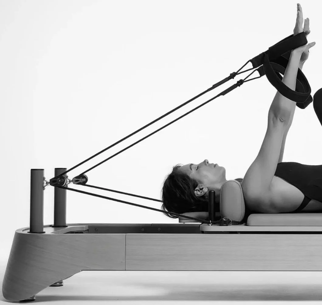 Woman exercising on a Pilates reformer machine, lying on her back with arms extended holding resistance bands.