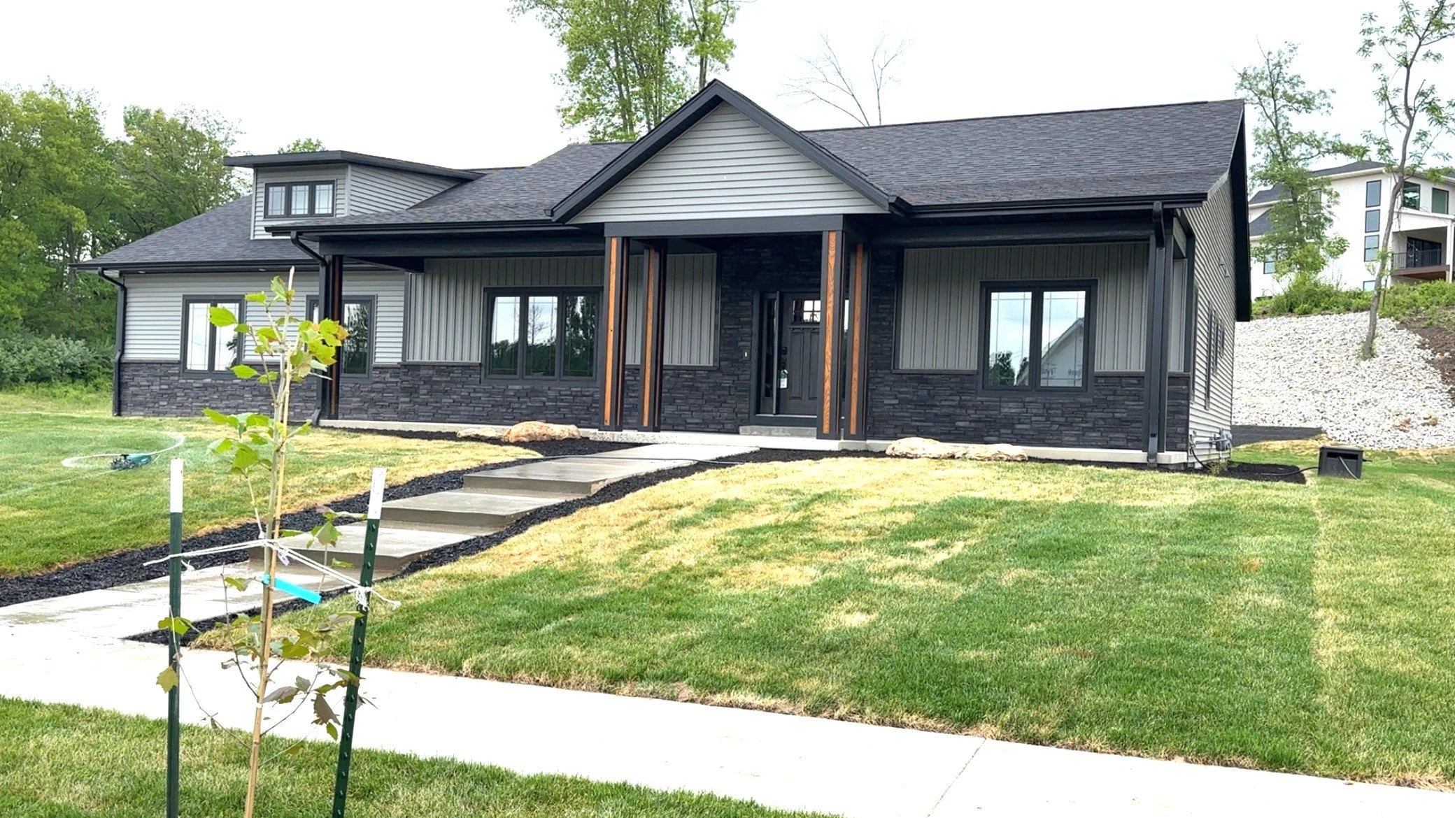 A modern house with dark stone and siding exterior, large windows, and a covered front porch, surrounded by a green lawn with a small tree and a concrete walkway leading to the entrance.