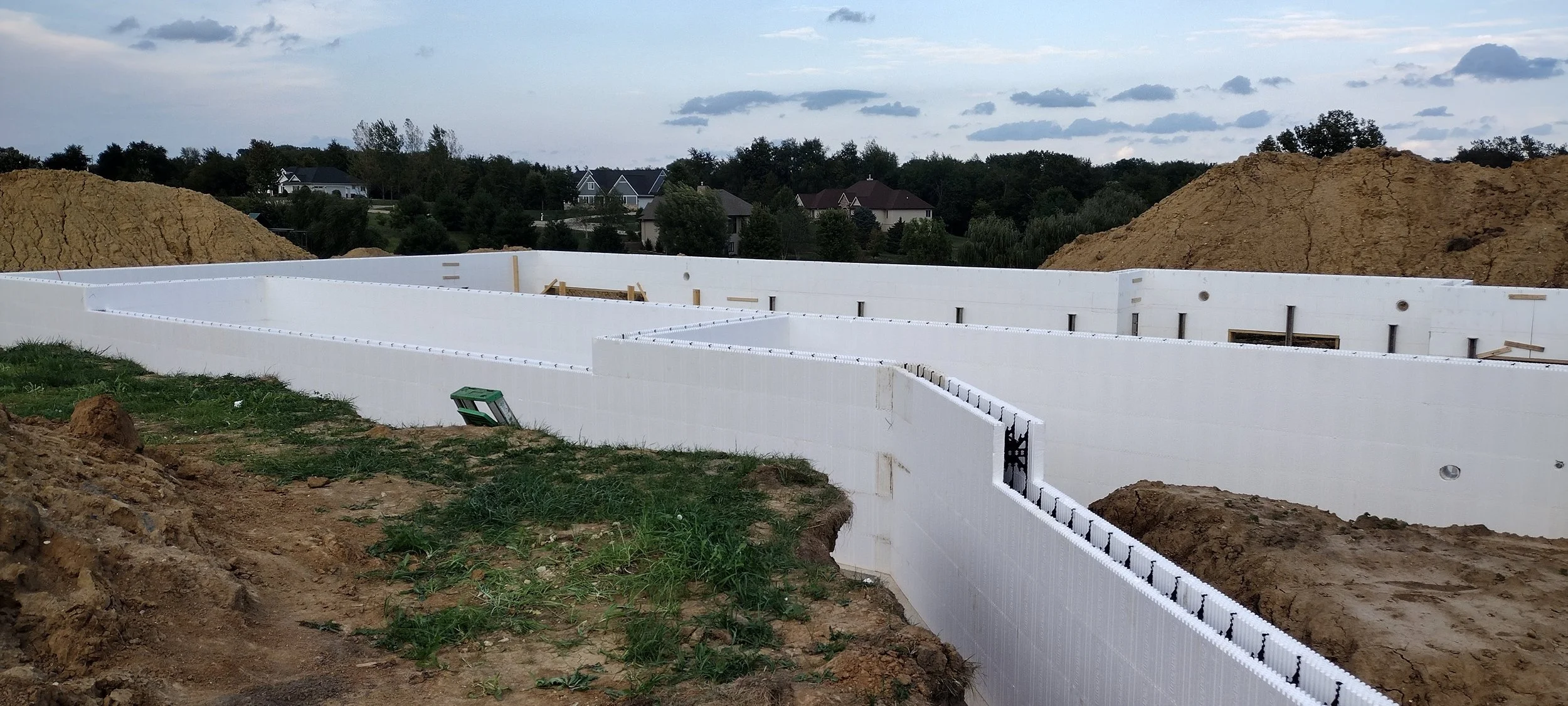 Construction site with a foundation made of insulated concrete forms (ICFs) in progress, surrounded by dirt piles and residential houses in the background.
