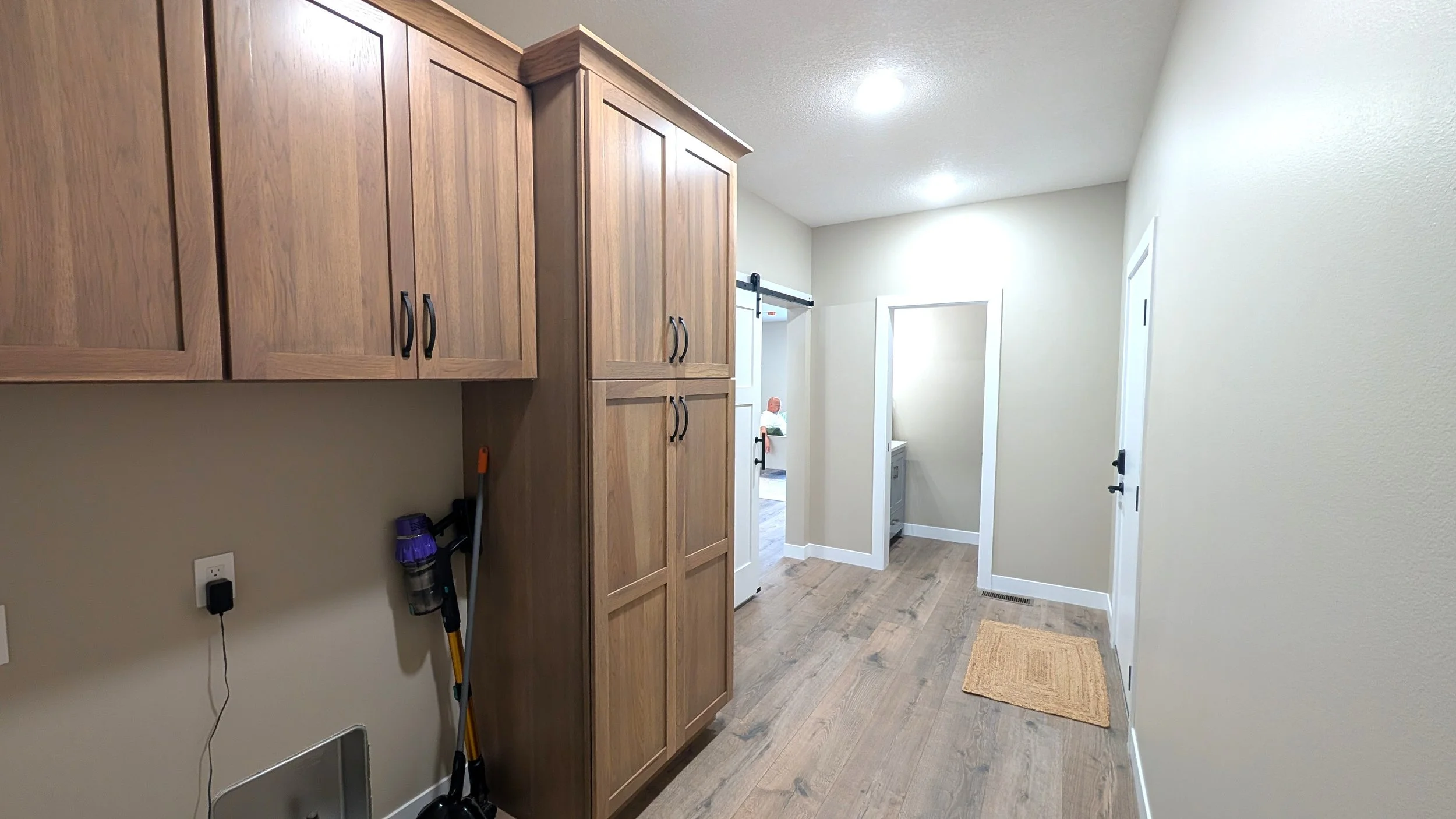 Interior view of a laundry room with beige walls, wooden cabinets, a small beige rug, and a doorway leading to a powder room.