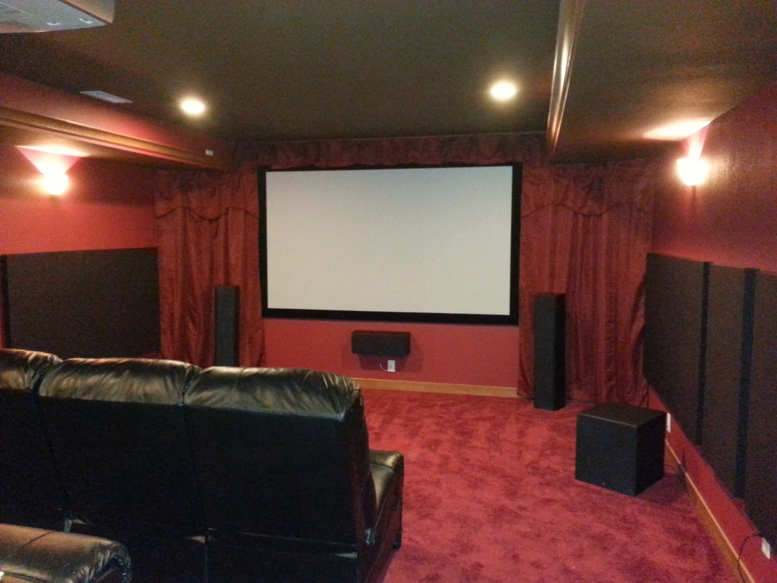 A home theater room with red walls, a large projection screen in the center, black leather couches, and Surround sound speakers.