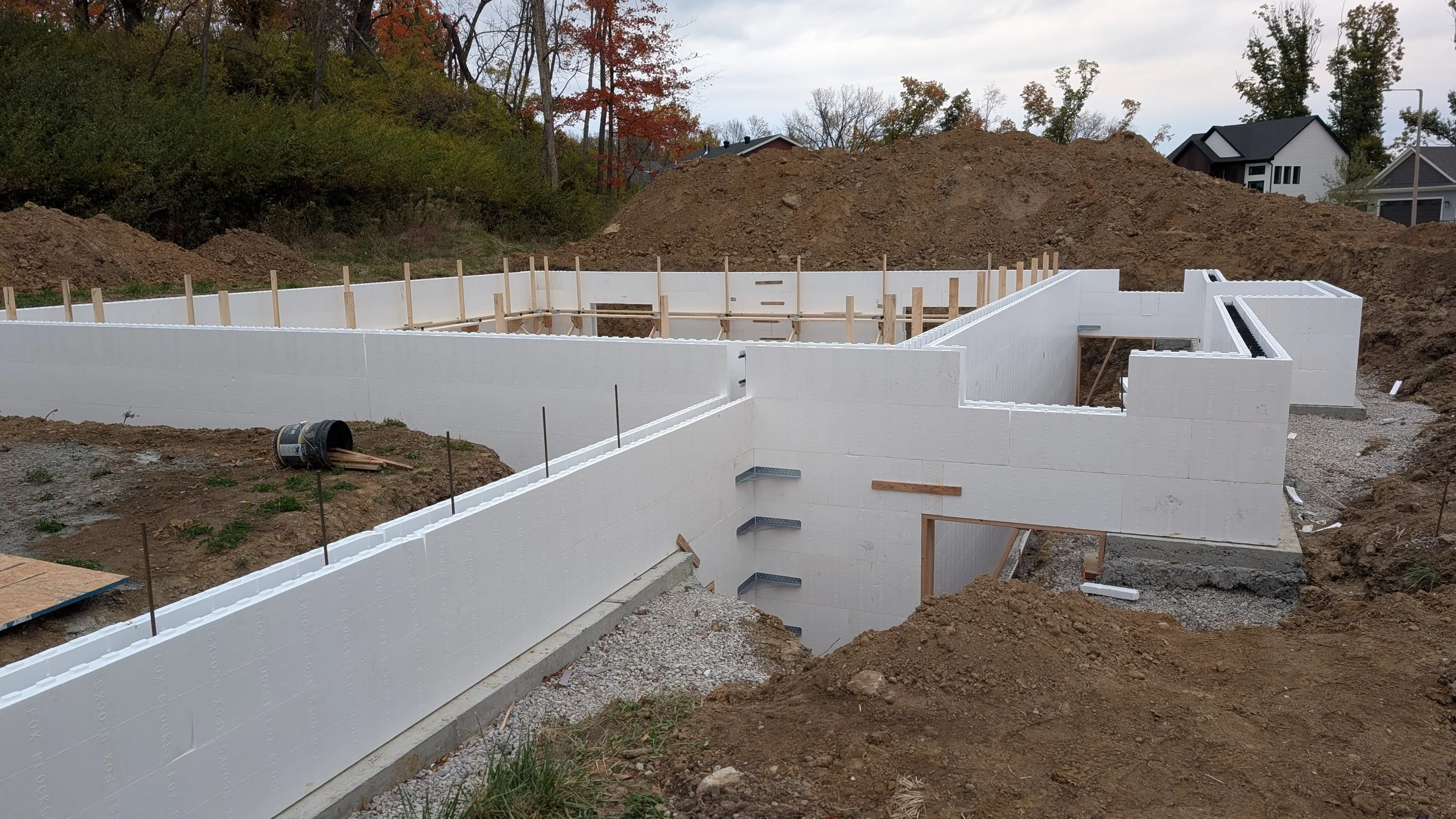Construction site with white insulated concrete forms being assembled for a building foundation, dirt mounds in the background, and neighboring houses visible behind the site.