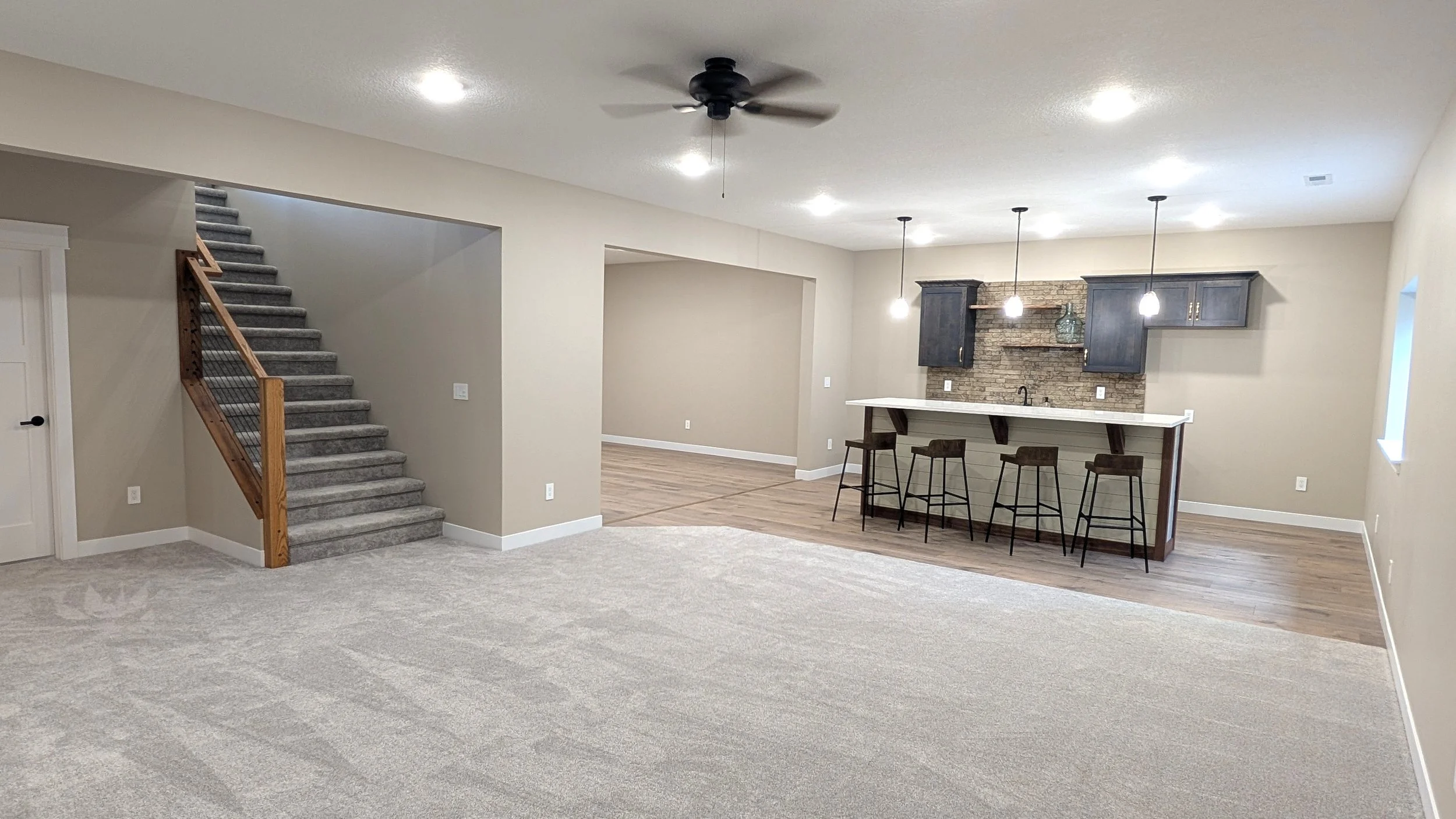 Empty open-concept living area with carpeted floor, stairway, wet bar with brick backsplash, dark cabinetry, and pendant lighting.