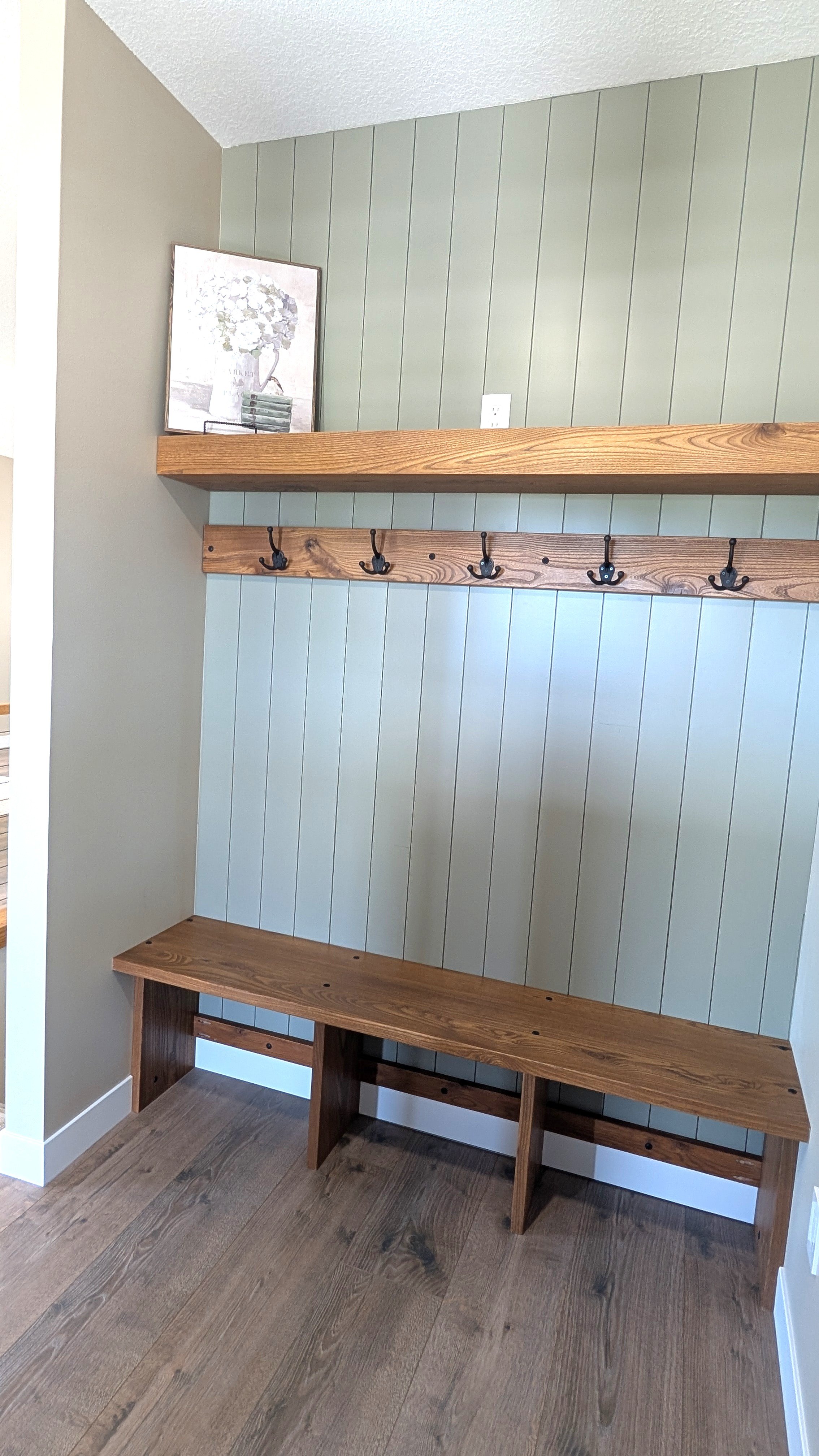A drop zone at the front door with a custom wood bench, and a shelf above with a framed picture of a vase with flowers and a stack of books. The floor is wooden, and there is a coat rack with hooks on the wall.