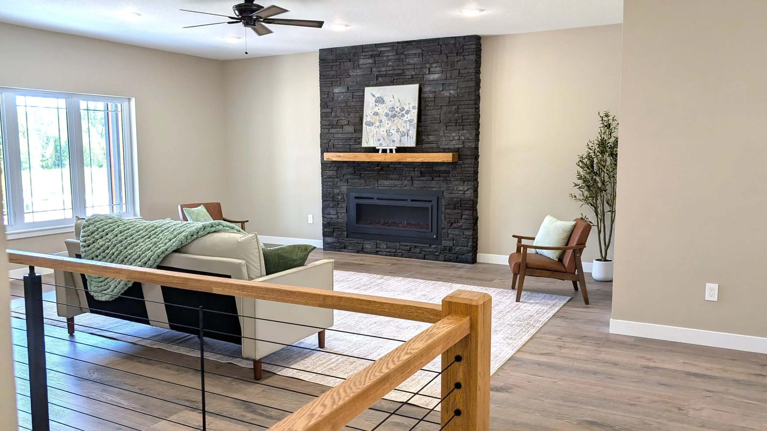 A living room with a black stone fireplace, a tan armchair, a white sofa with a green blanket, and large window with white blinds.